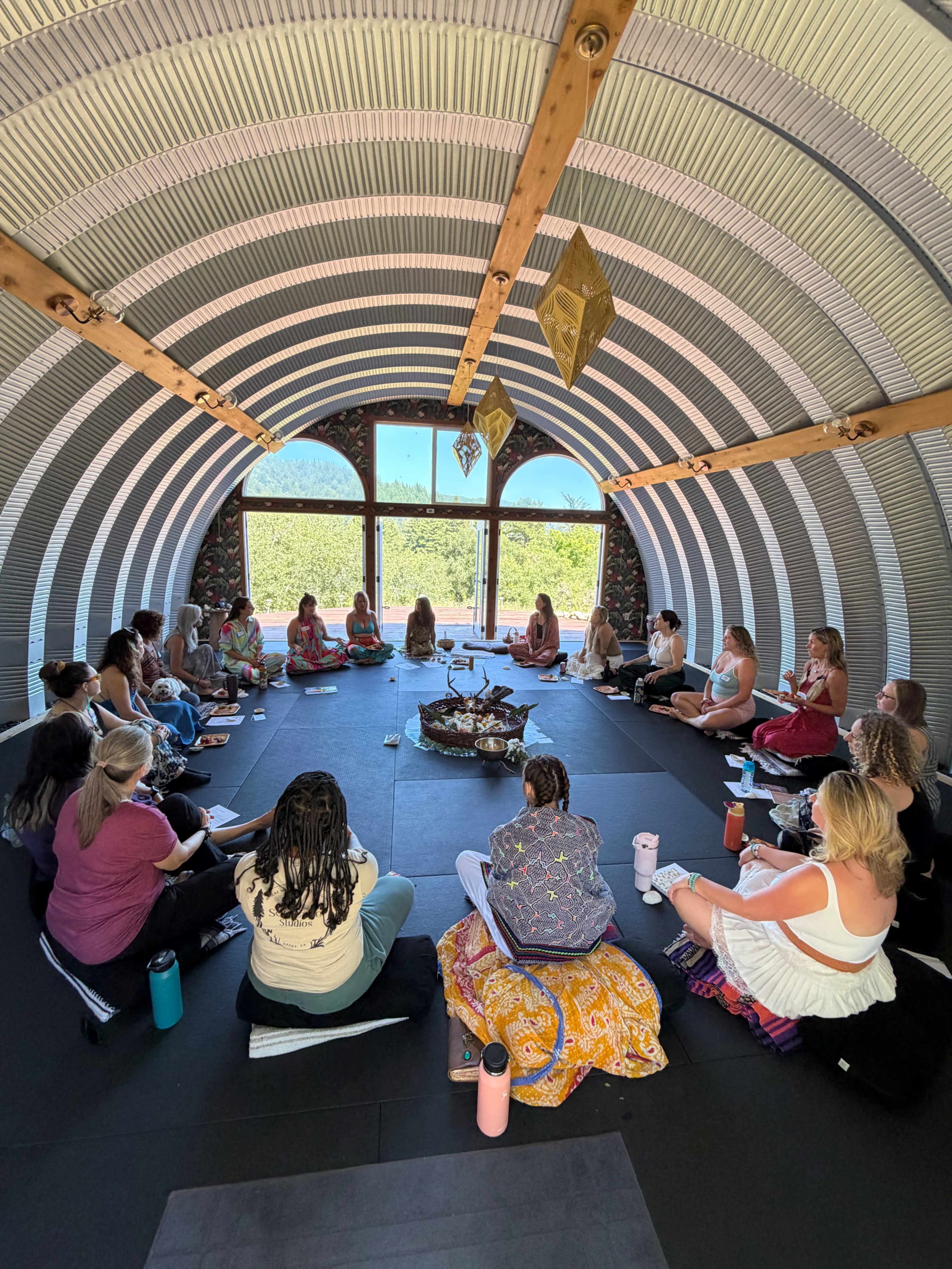 A group of people is seated in a semicircle on the floor of a large, curved indoor space with windows overlooking greenery, engaged in a gathering centered around a decorative focal point.
