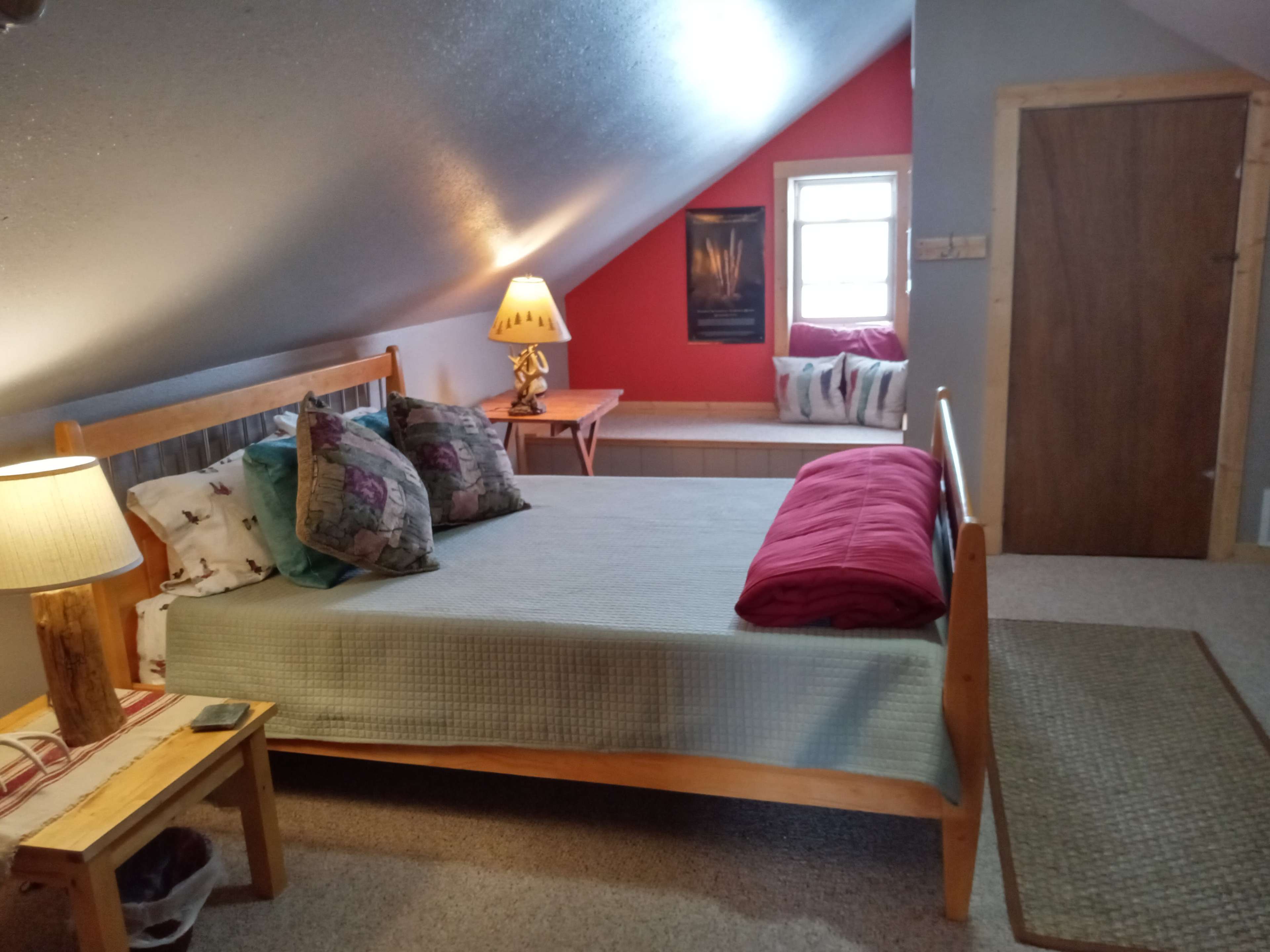 The image shows a cozy attic bedroom featuring a wooden bed with decorative pillows, a lamp on a side table, and a red accent wall.