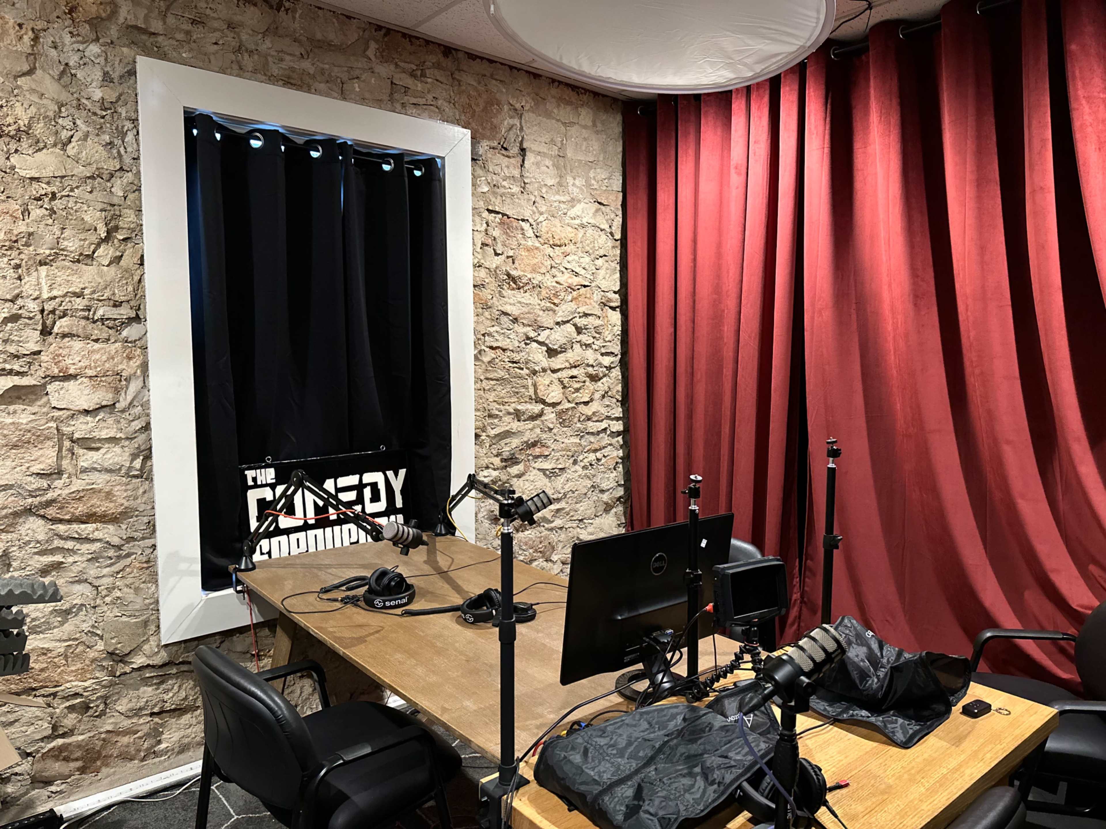 The image shows a recording studio with a wooden table, several microphones on stands, and a backdrop of red curtains next to a stone wall.