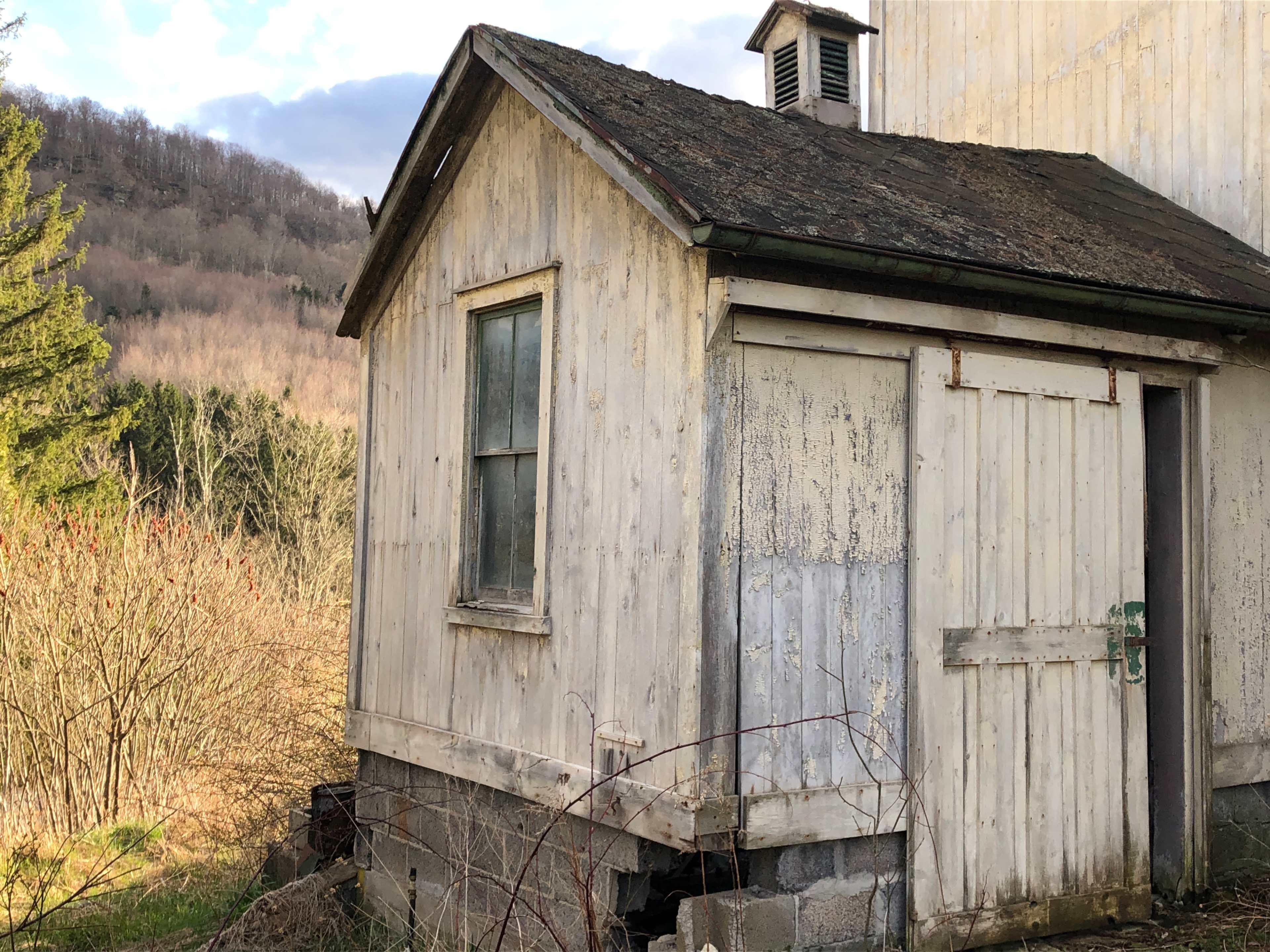 The image shows an aged wooden shed with a partially open door, situated beside a hill with sparse vegetation.