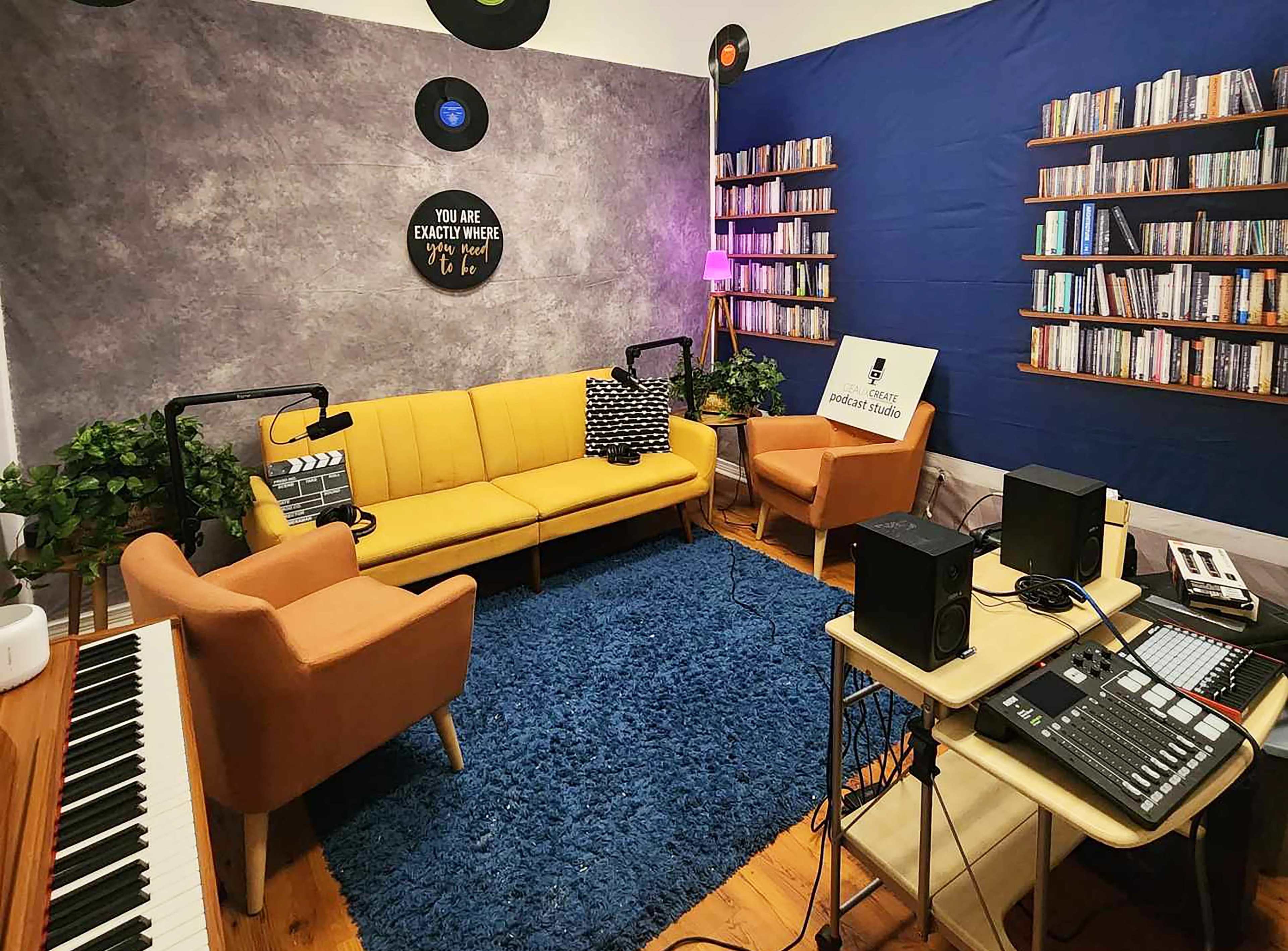 The image shows a music studio featuring a yellow sofa, two armchairs, a blue wall with bookshelves, and various audio equipment on a desk.
