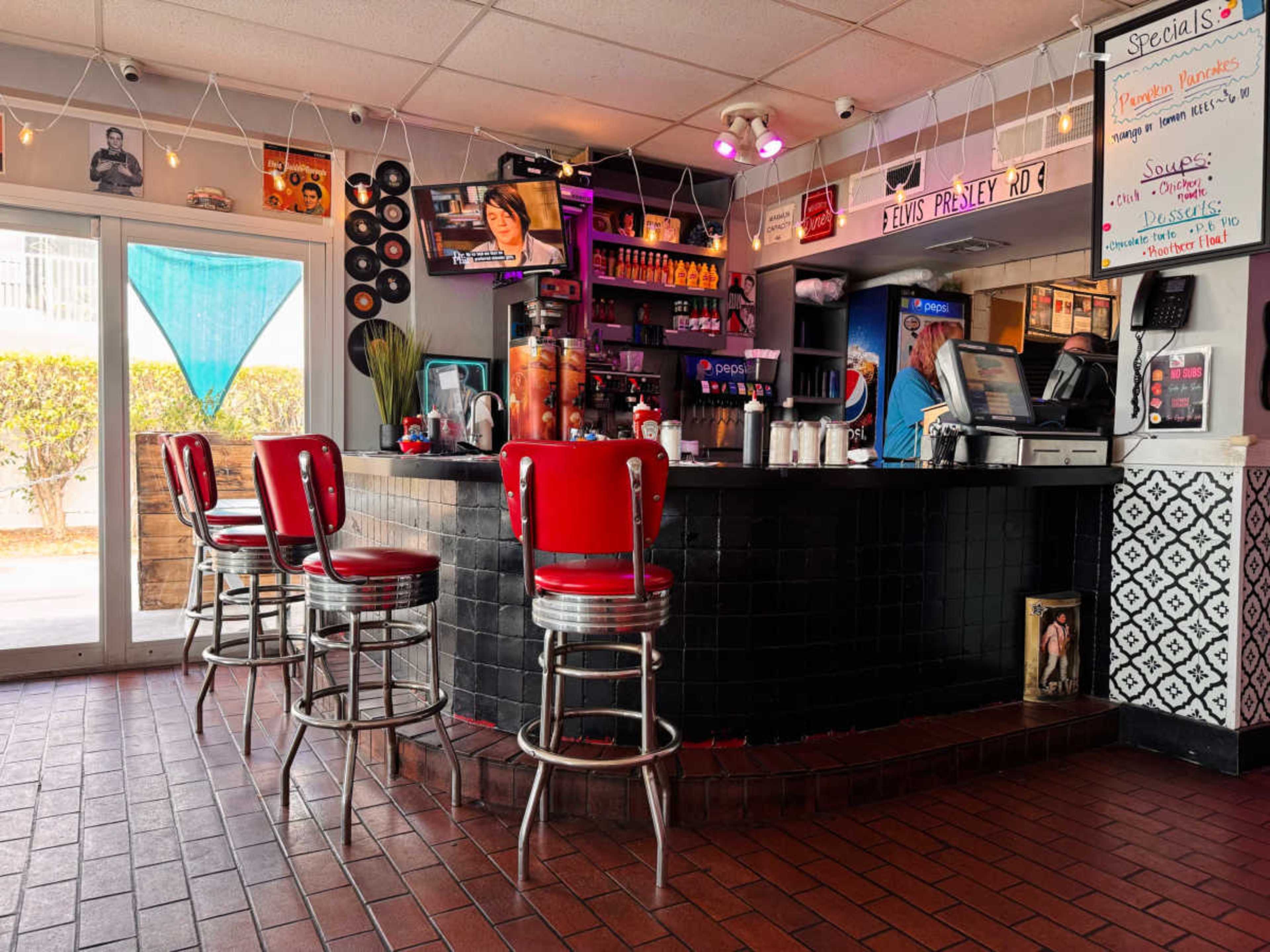 The image shows a diner-style bar with red leather stools, a black countertop, and a menu board featuring daily specials, located near a glass door leading outside.