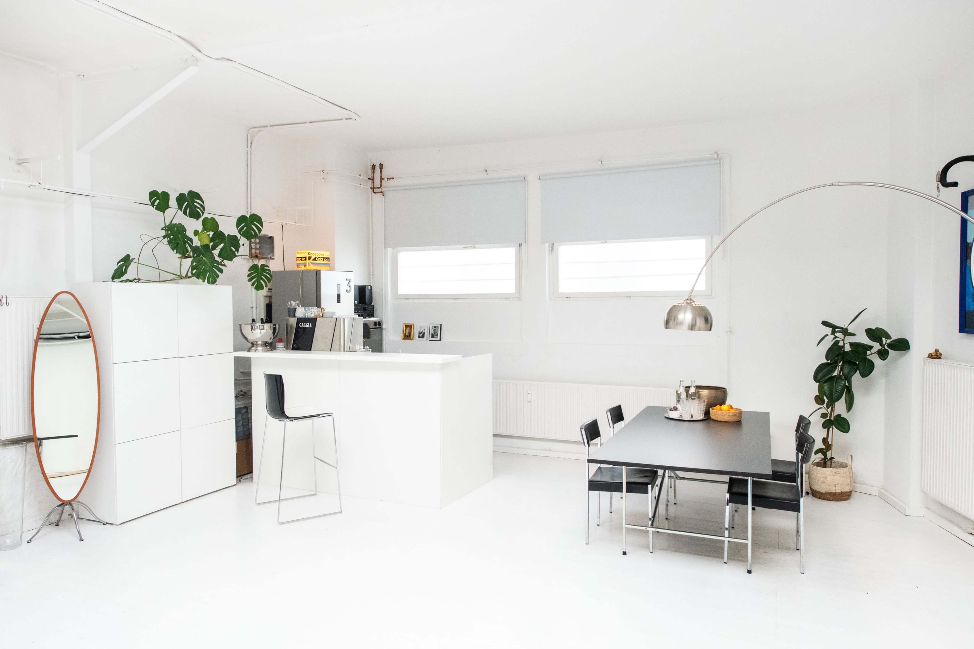 The image shows a modern, minimalist kitchen and dining area with white walls, a large window, and simple furnishings.