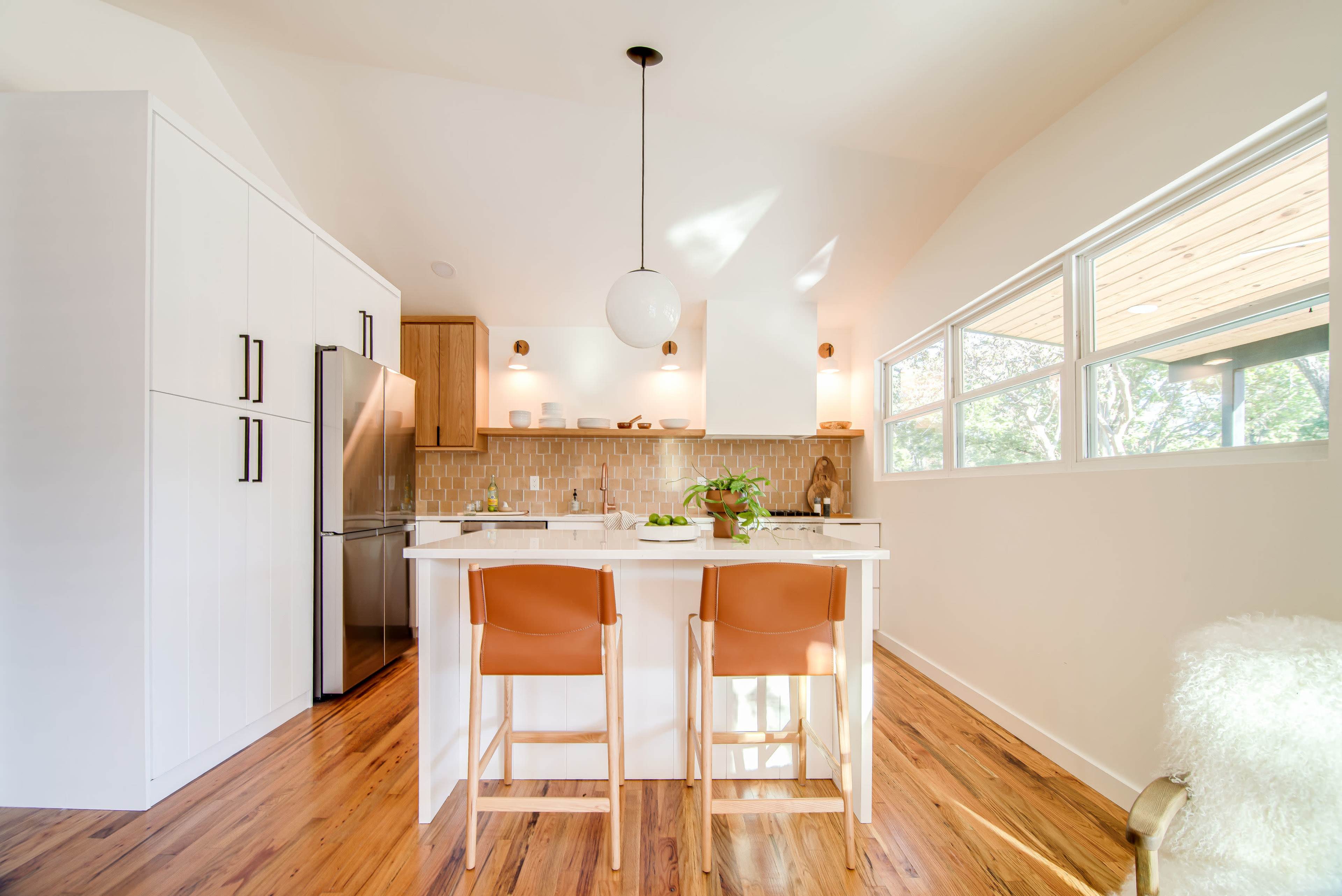 A modern kitchen features a white island with two tan chairs, a stainless steel refrigerator, and large windows that allow natural light to illuminate the space.