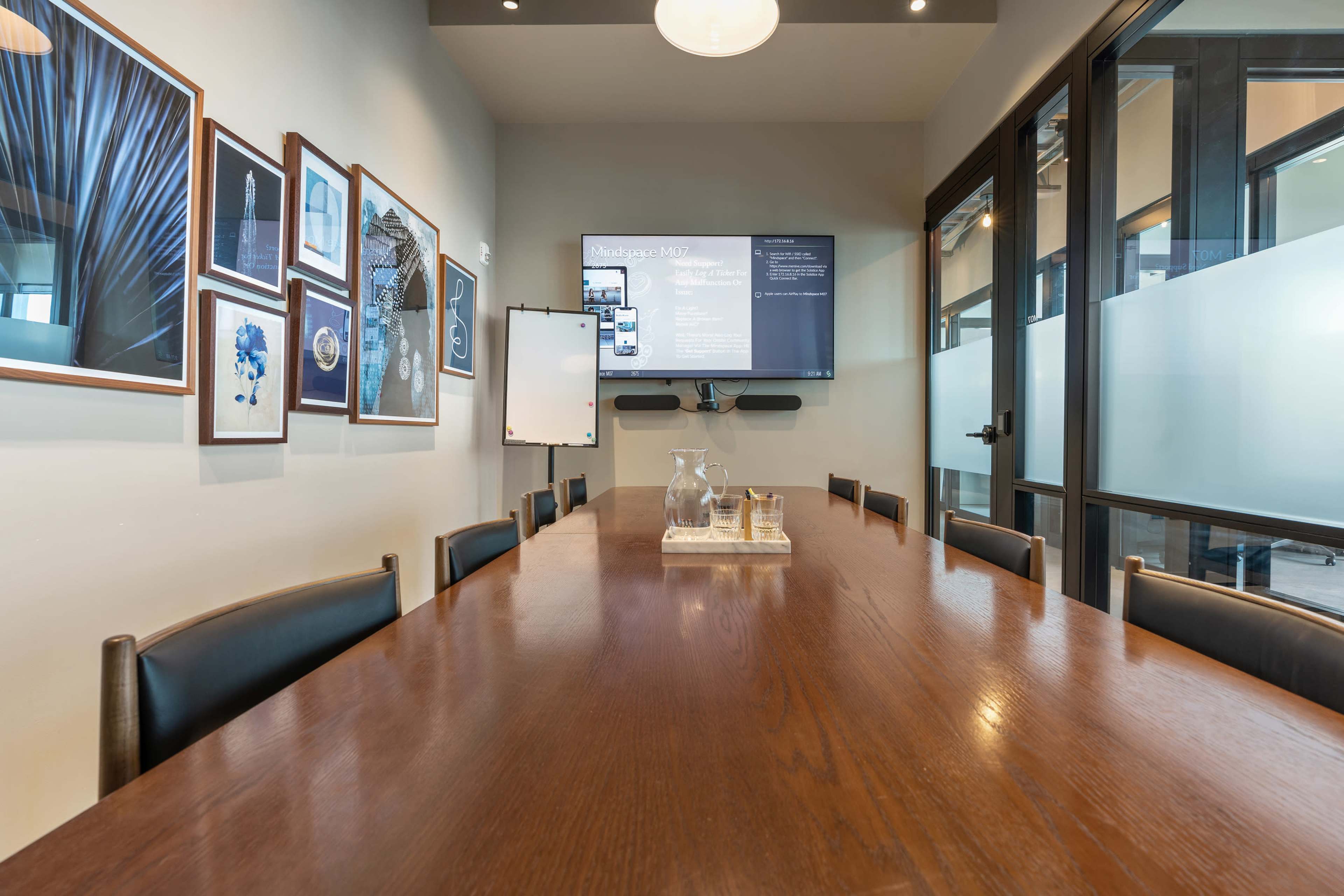 A conference room with a long wooden table, black leather chairs, a television display, and framed art on the walls.