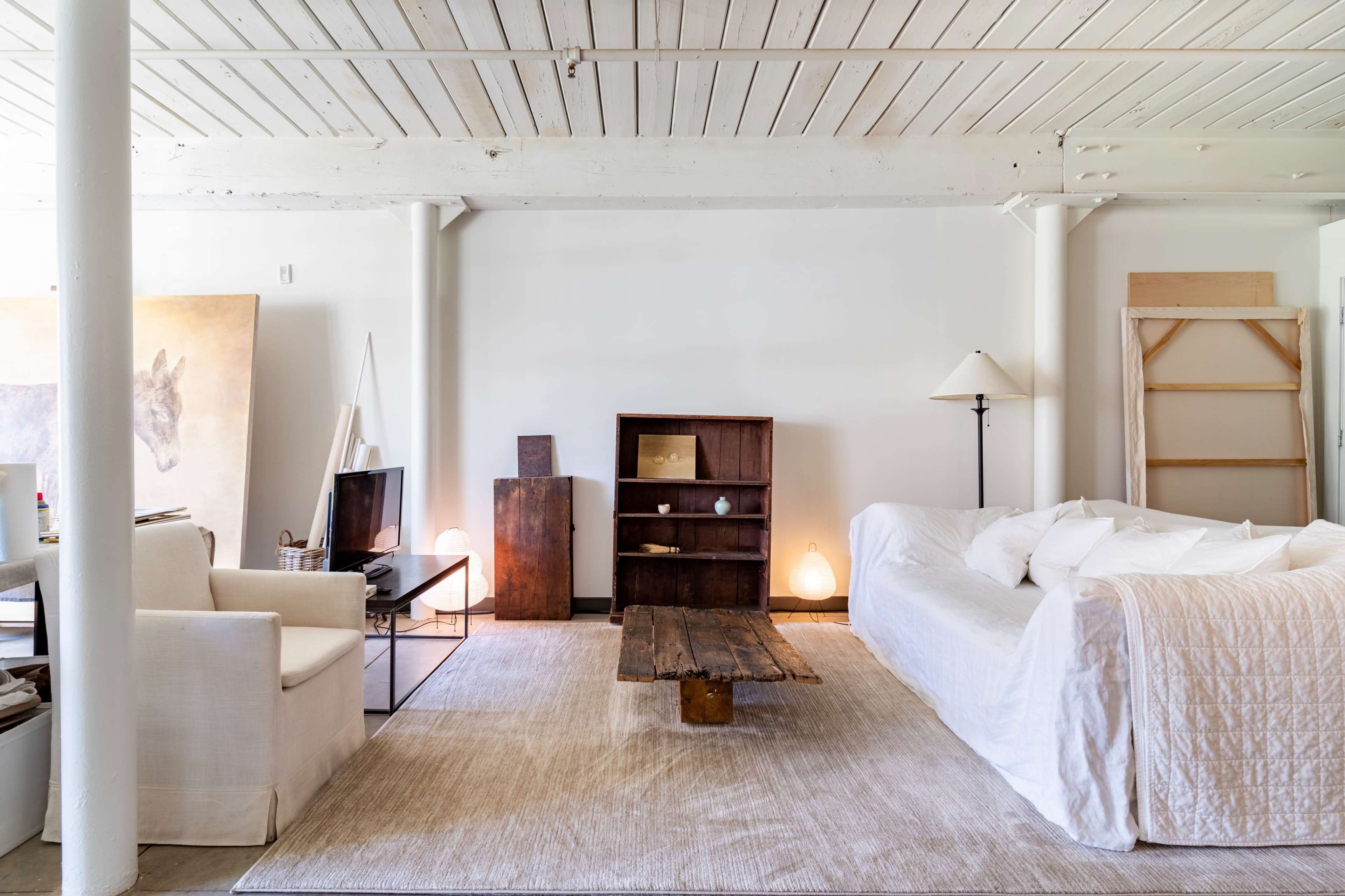 A spacious living room features a white couch, a wooden coffee table, a television, and minimalist decor against a backdrop of light-colored walls and wooden beams.