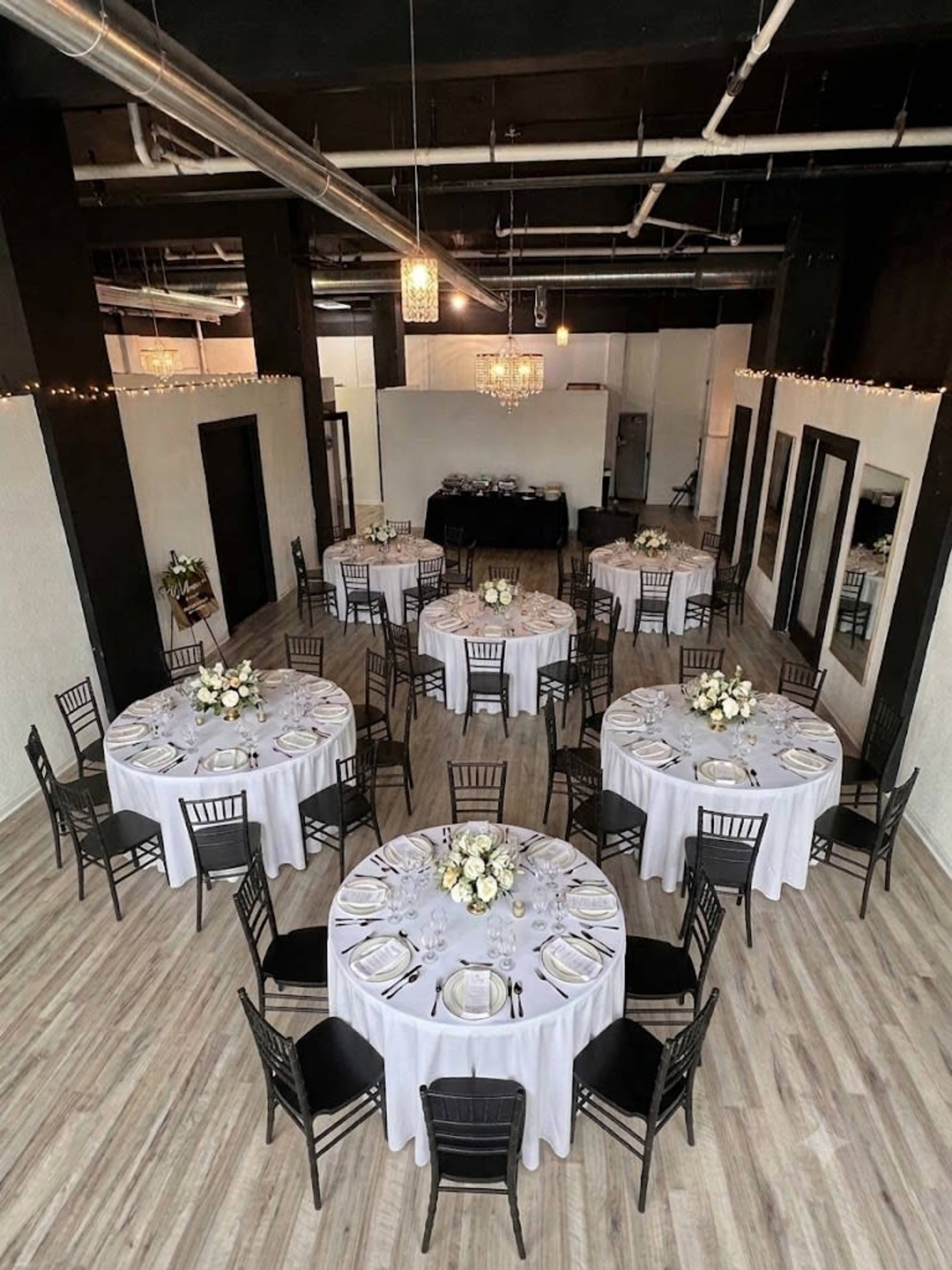 A spacious event venue features multiple round tables set for dining, each adorned with white tablecloths and floral centerpieces.