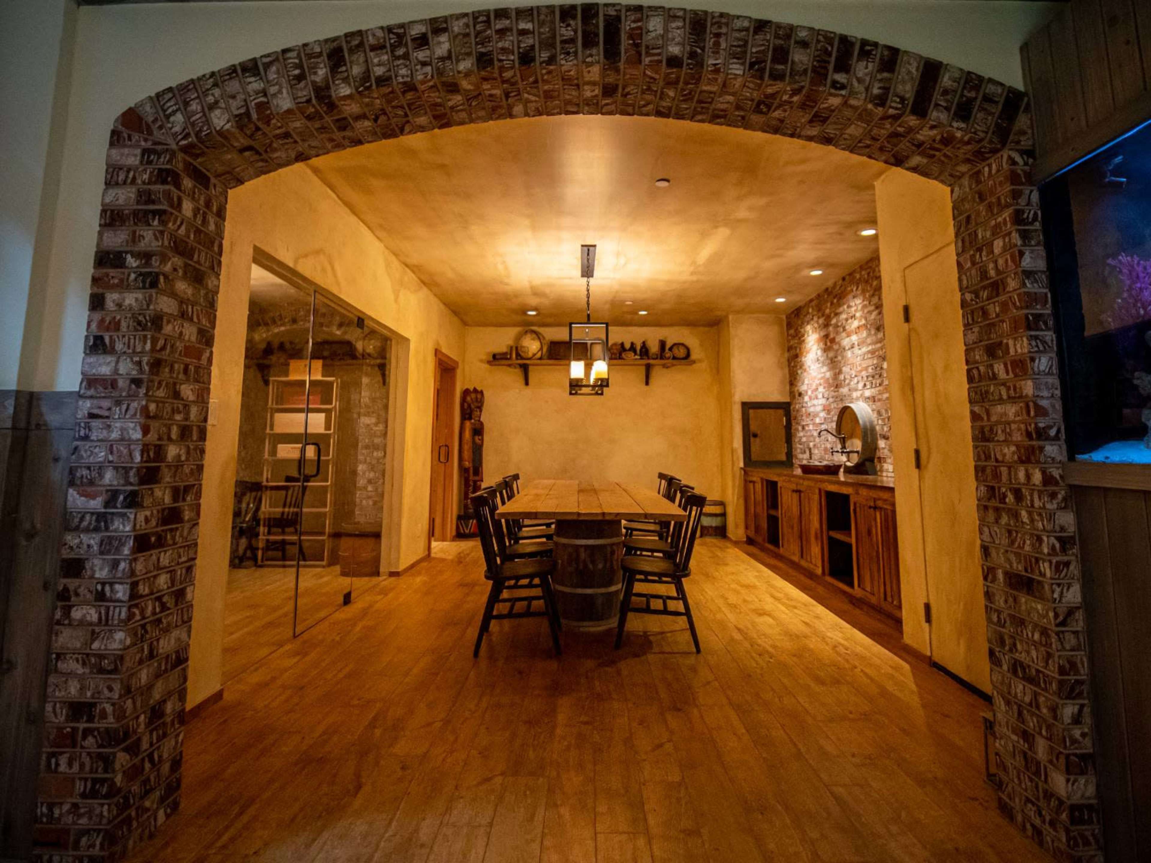The image shows a warm, rustic dining room with wooden flooring, a long table surrounded by chairs, and a brick archway leading to another area.