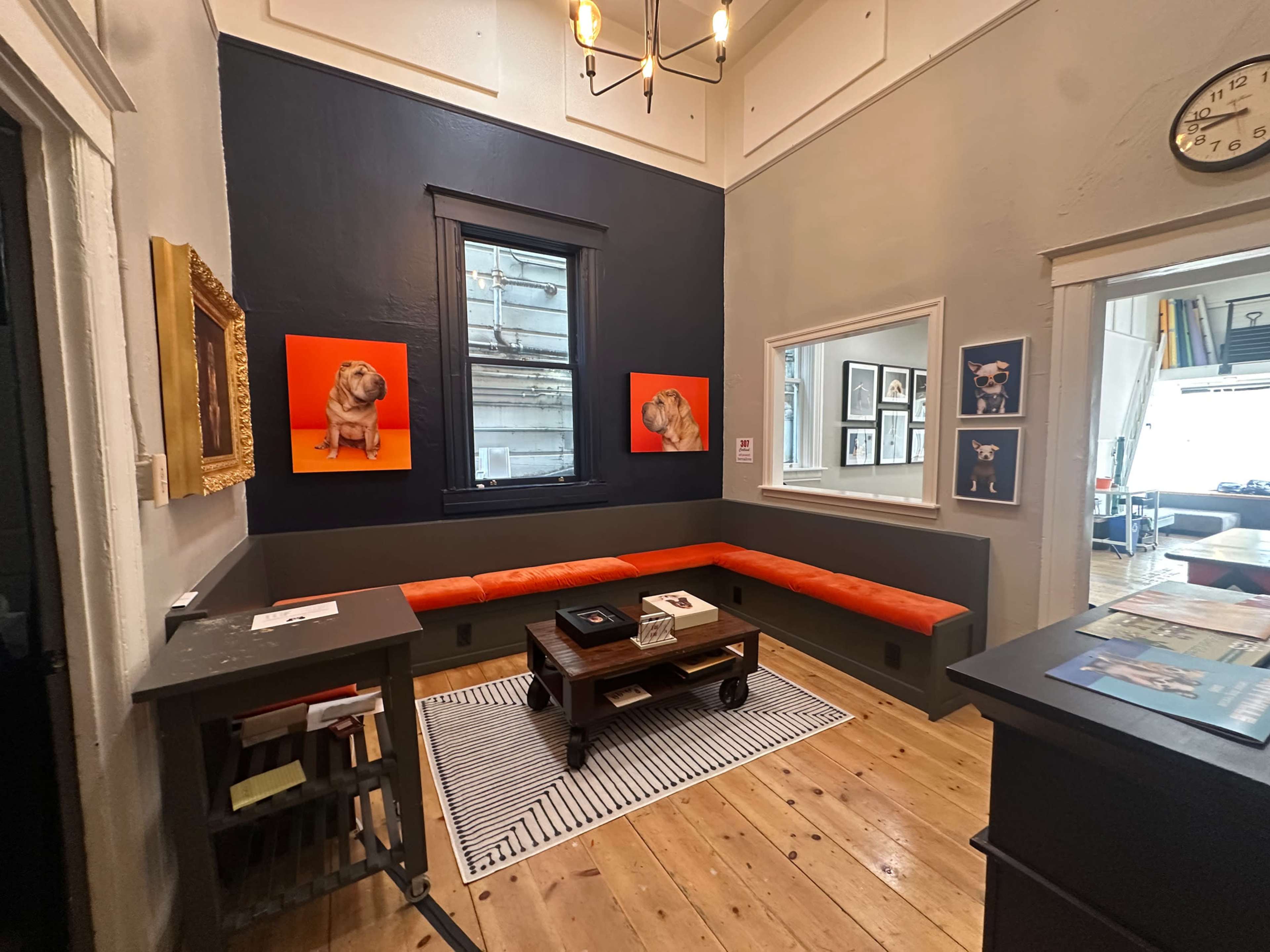 The image shows a small room with a wooden floor, featuring an orange bench along the wall, framed dog portraits, and a table with a magazine display near a window.