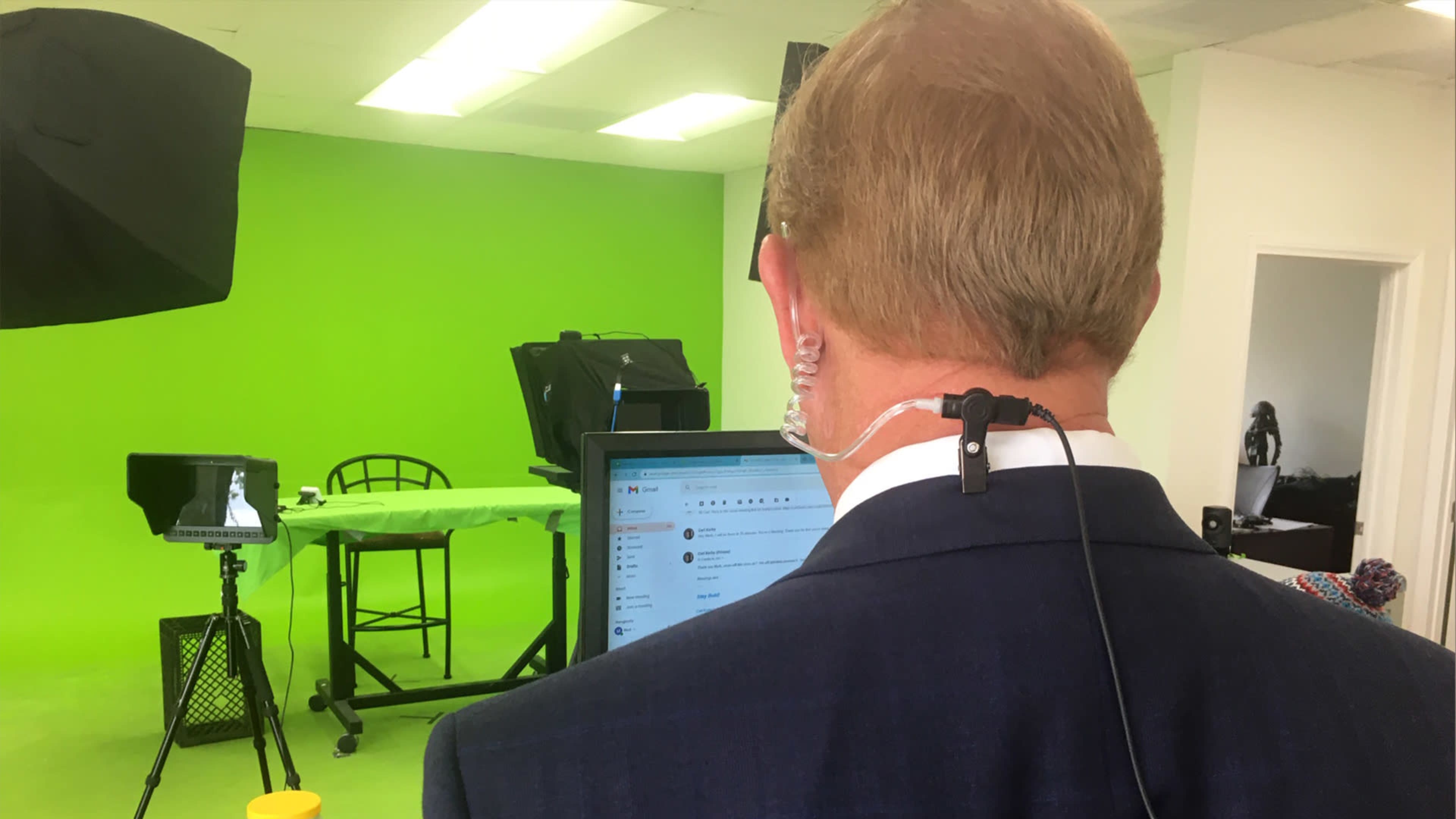 A person in a suit stands with their back to the camera in a studio featuring a green backdrop and various filming equipment.