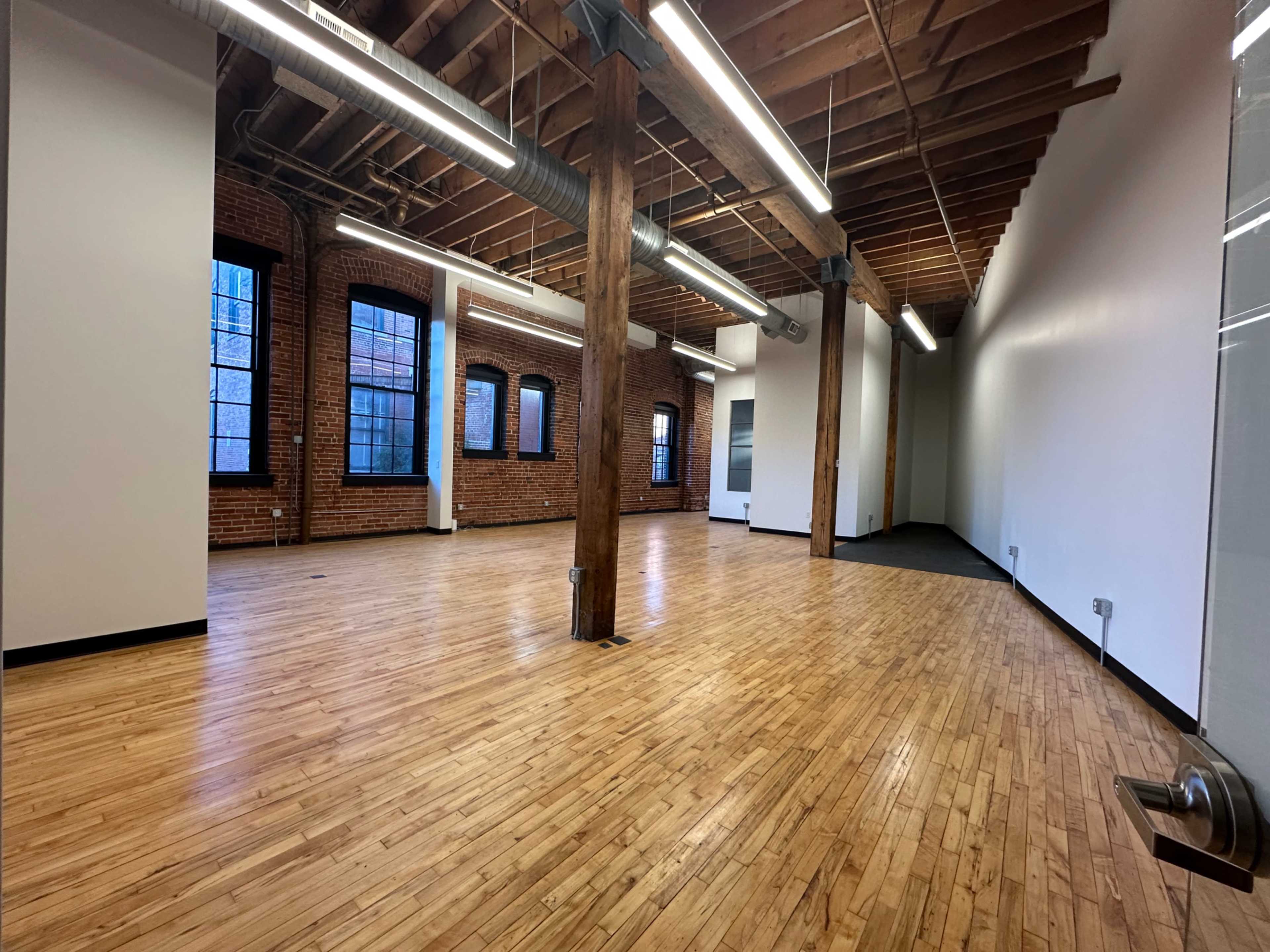 The image shows an empty office space with hardwood floors, exposed brick walls, and wooden beams supporting the ceiling.