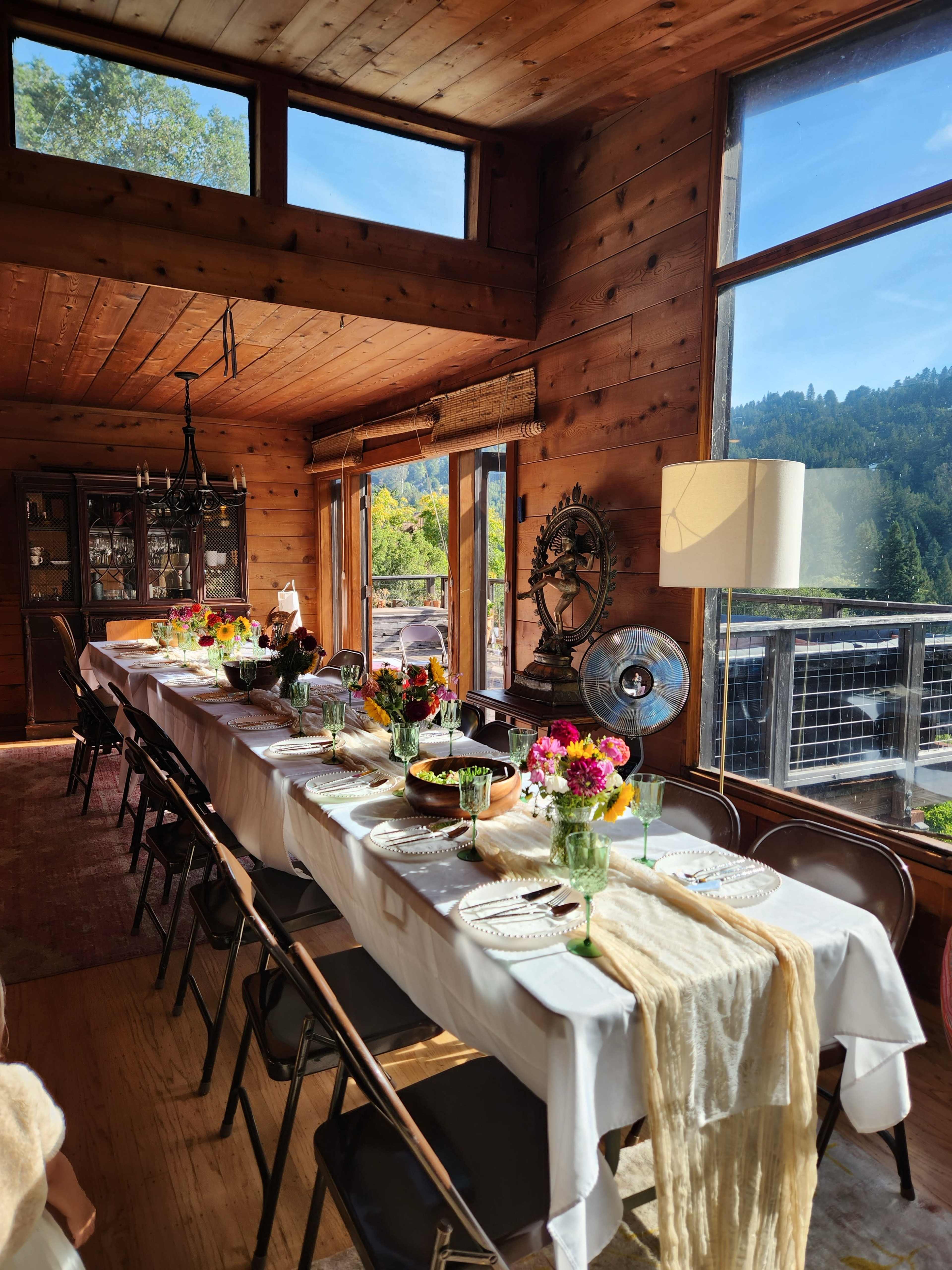 A long dining table is set with white tablecloths and decorative centerpieces in a sunlit wooden room featuring large windows and a view of the mountains.