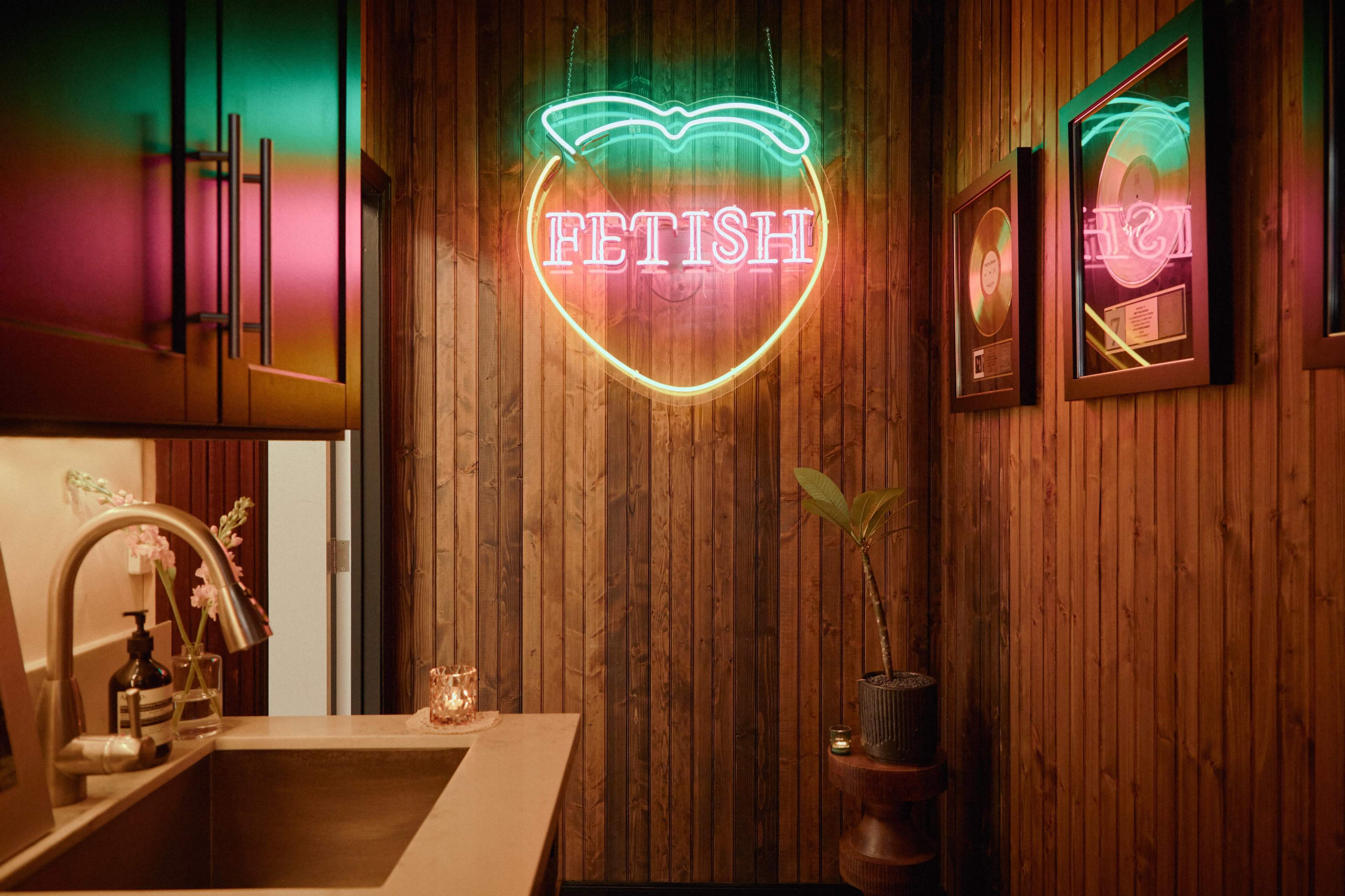 A narrow hallway features a neon sign that reads "FETISH" alongside framed records and a small plant near a sink.