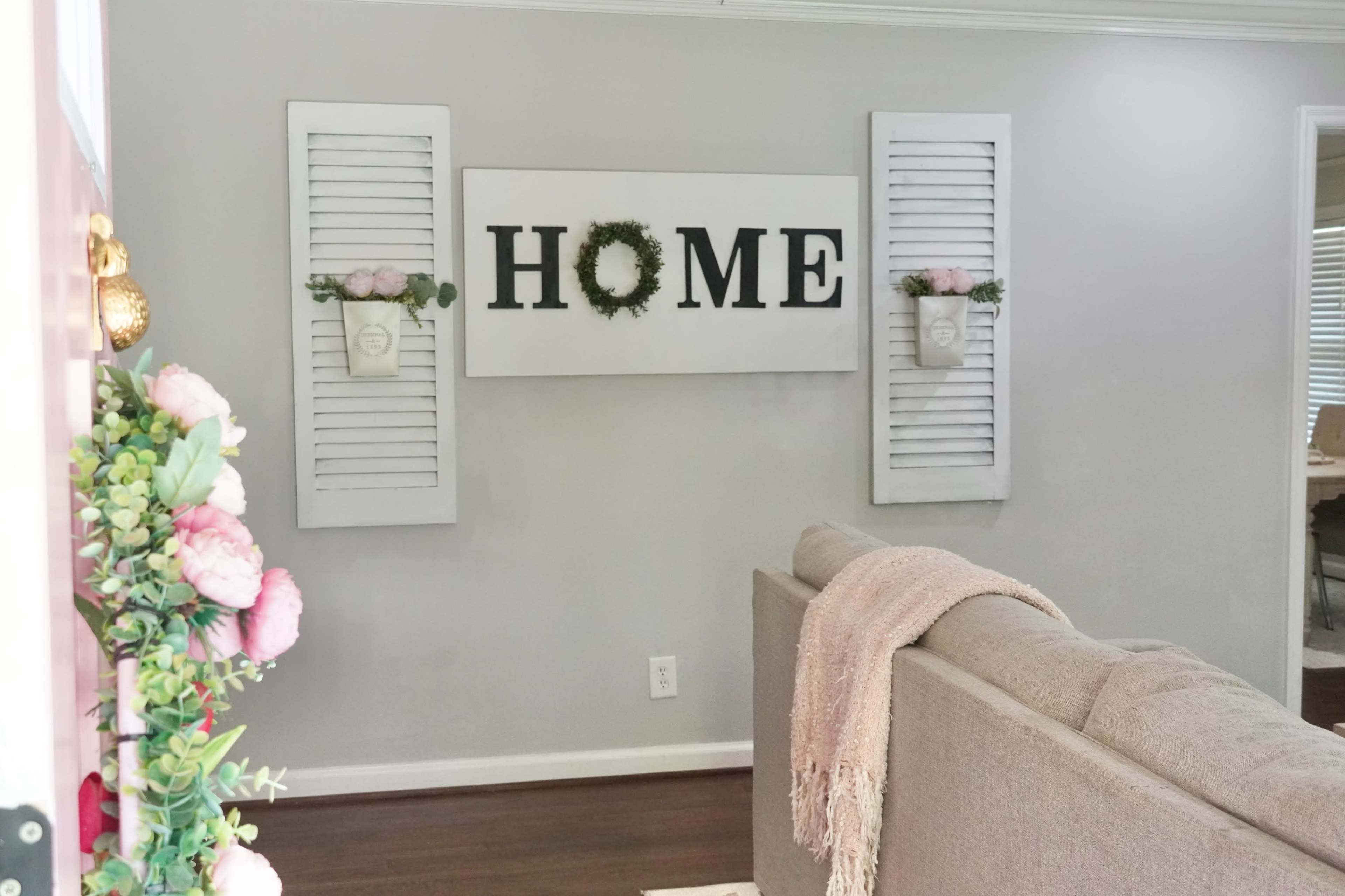 A cozy living room features a large wall art piece that reads "HOME" flanked by decorative shutters and planters with floral accents.