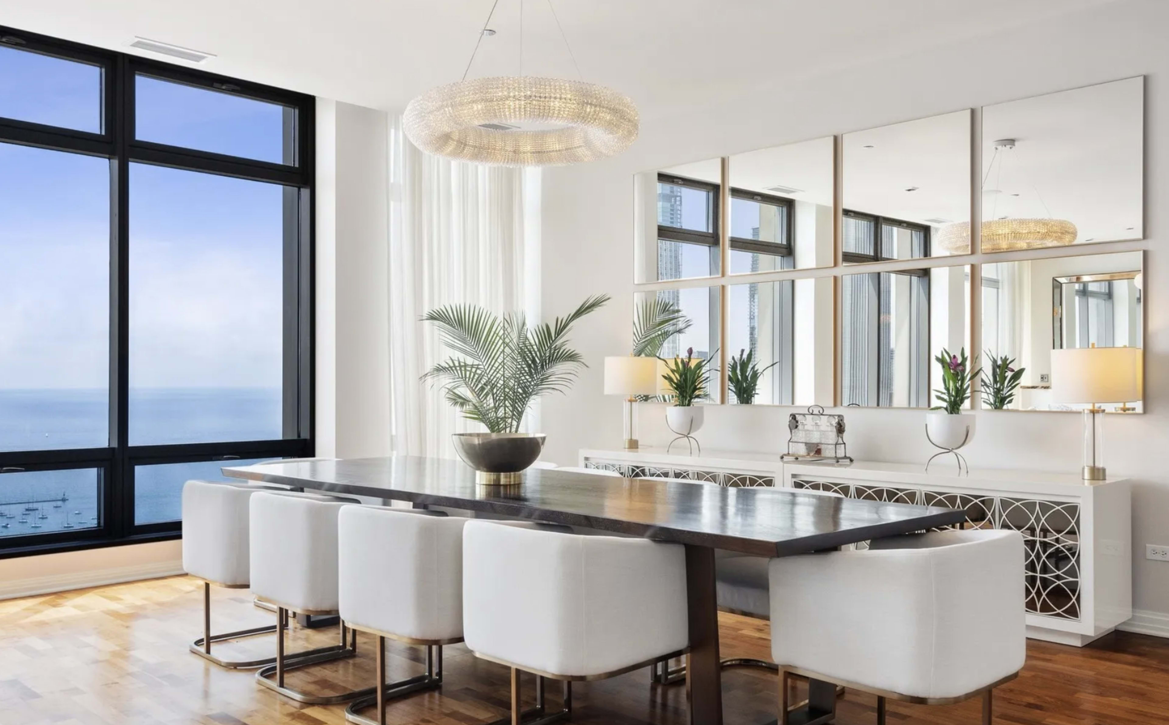 The image depicts a modern dining area with a large wooden table surrounded by upholstered chairs, large windows offering a view of the sea, and decorative elements including a chandelier and mirrored panels on the wall.