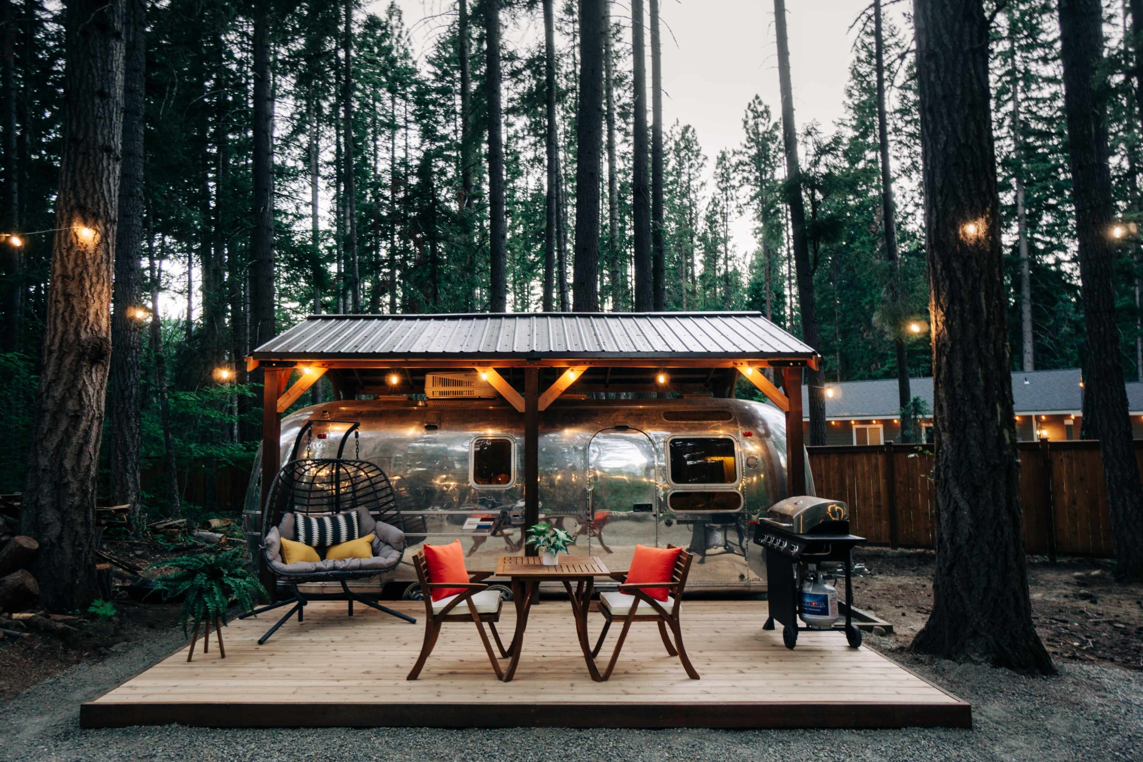 A shiny Airstream trailer is set up under a wooden gazebo in a forested area, featuring a seating area with a table and chairs, a grill, and hanging string lights.