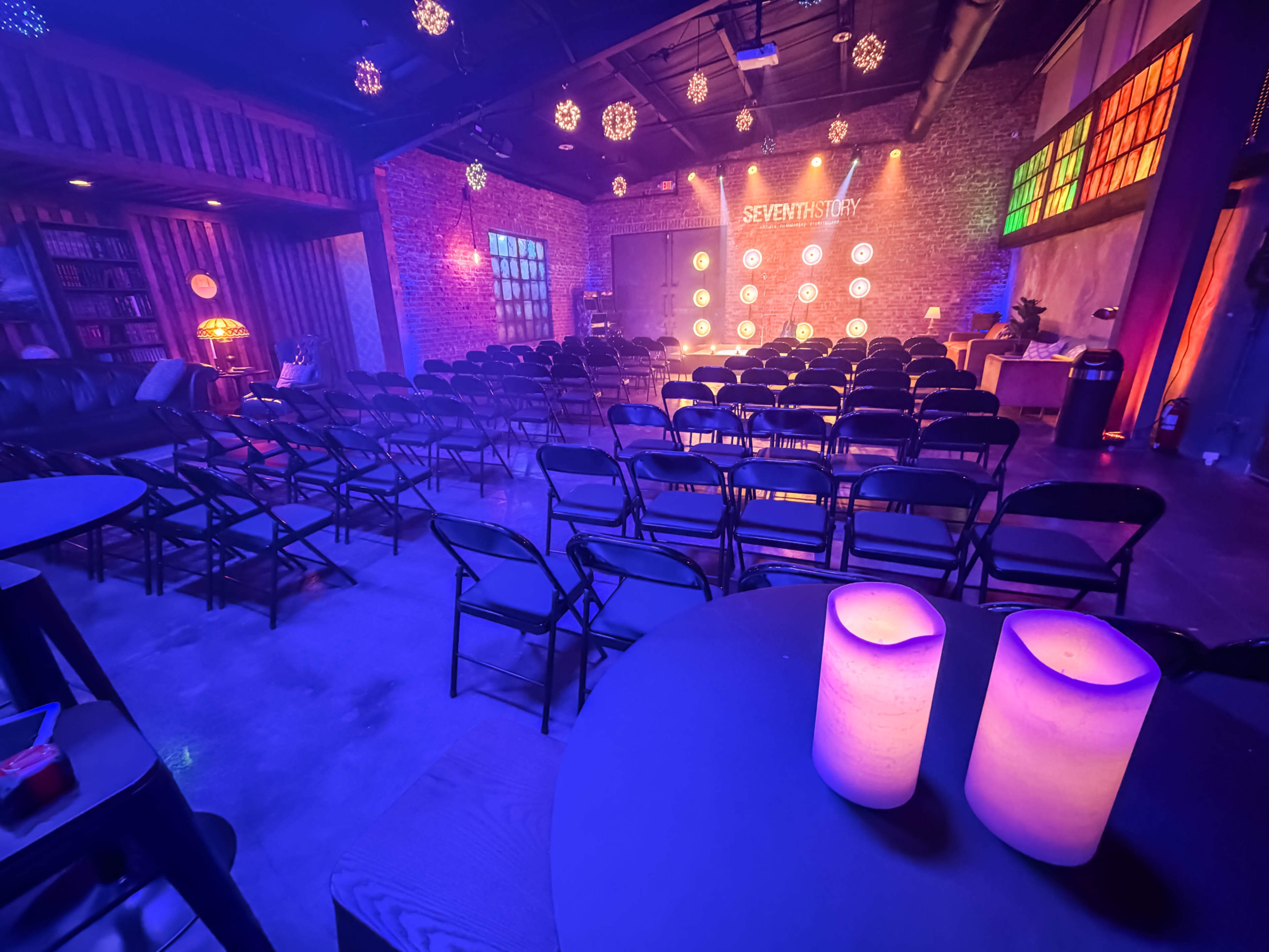 The image shows a dimly lit venue with rows of black chairs set up in front of a brick wall featuring circular light fixtures and decorative elements.
