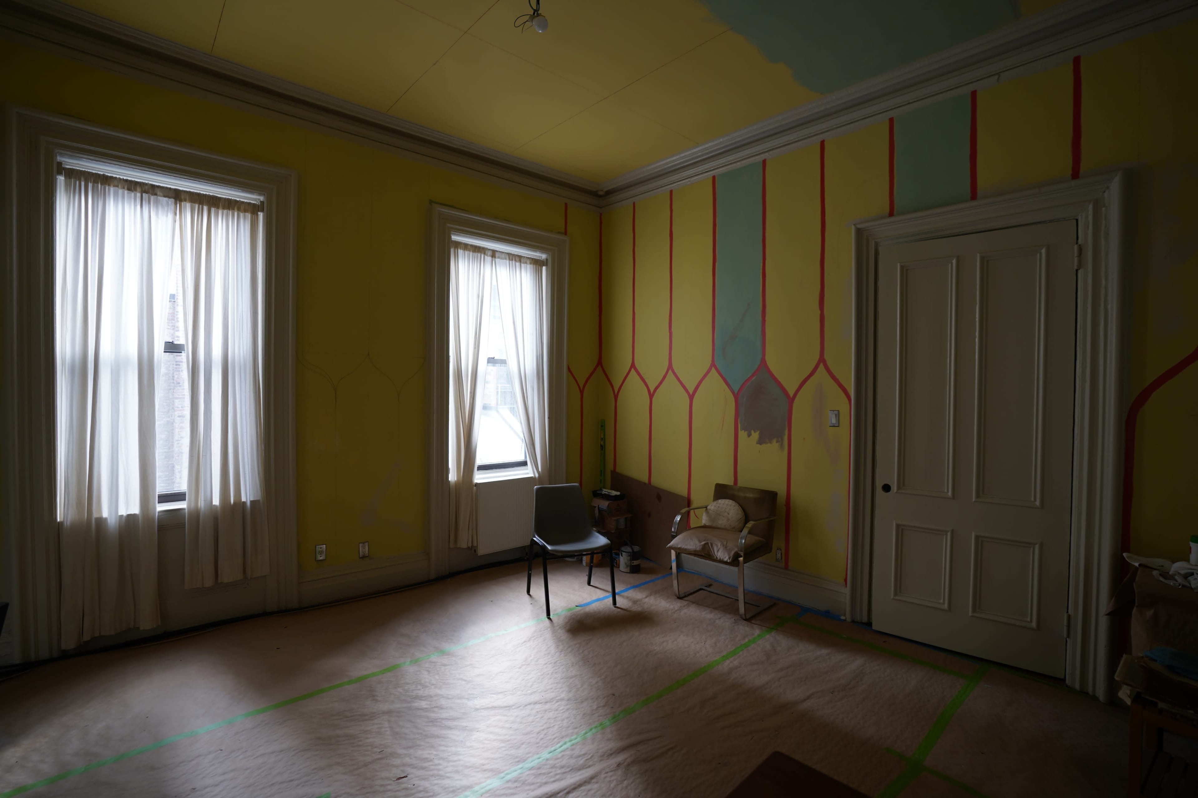 A room with yellow walls, two windows covered by sheer curtains, and two chairs positioned in the corner on a protected floor.