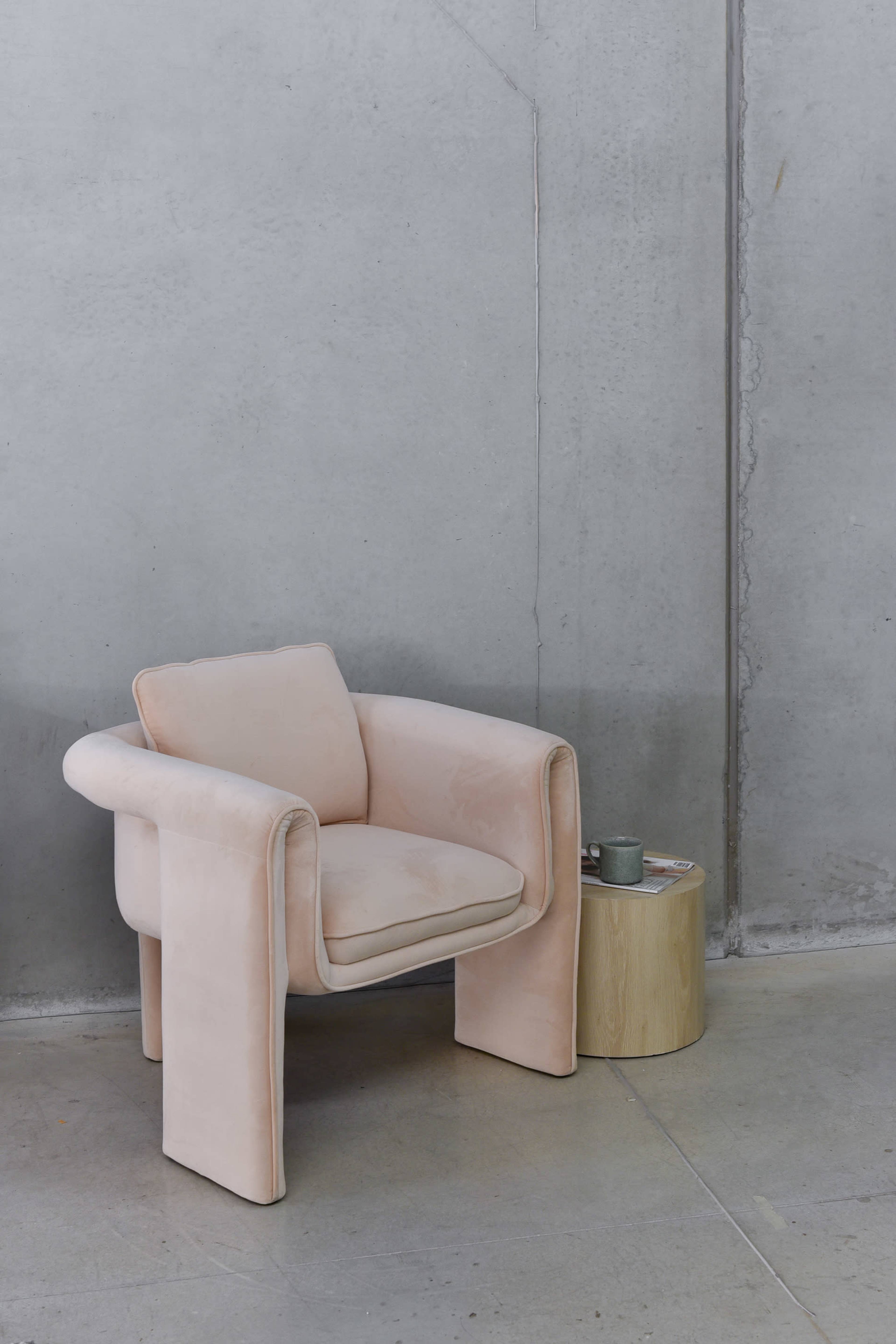 A pink upholstered chair is positioned next to a small wooden side table in a minimalist, concrete room.