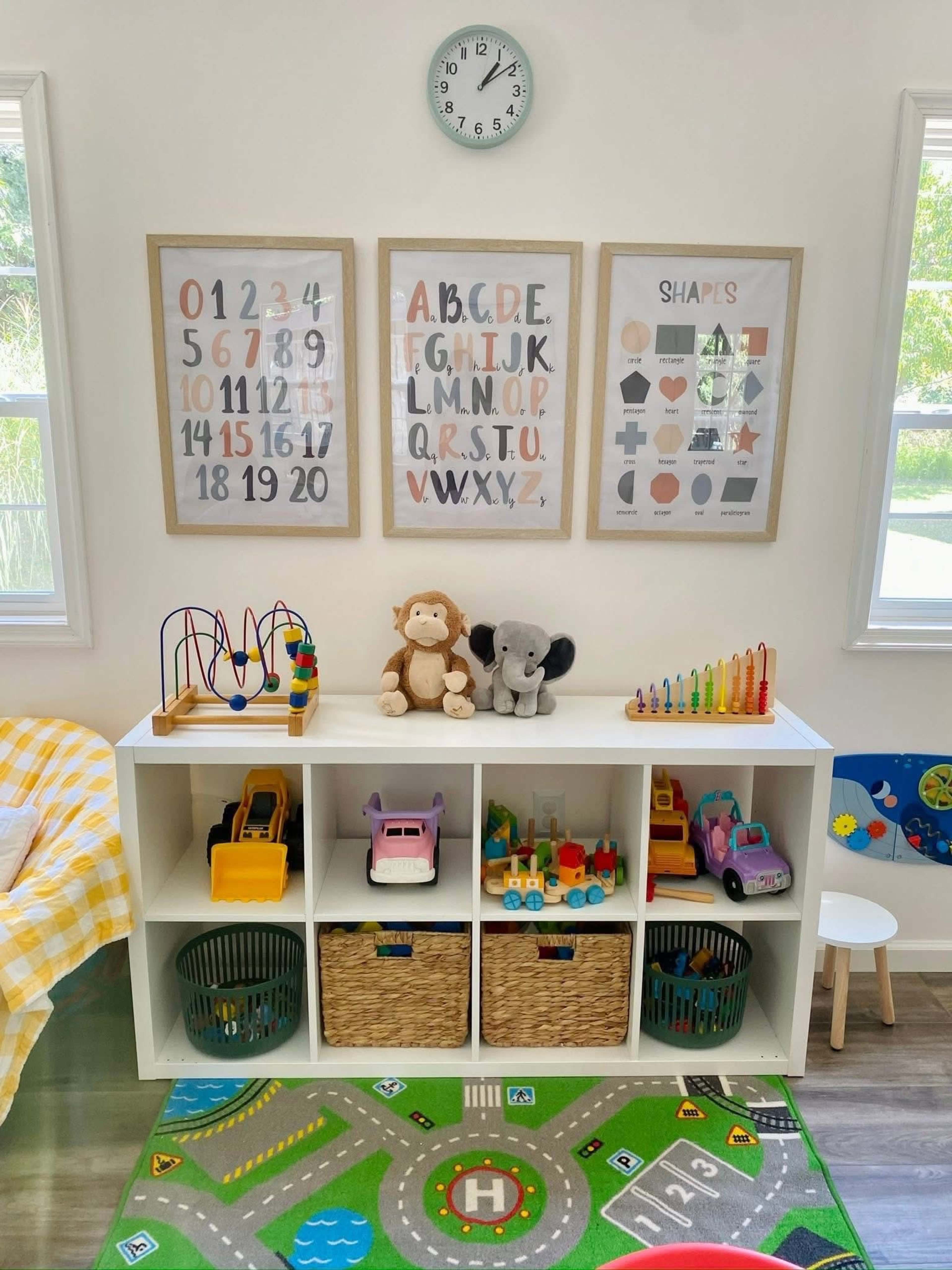 The image shows a playroom with a white storage unit filled with toys, accompanied by framed educational posters on the wall and a colorful play mat on the floor.
