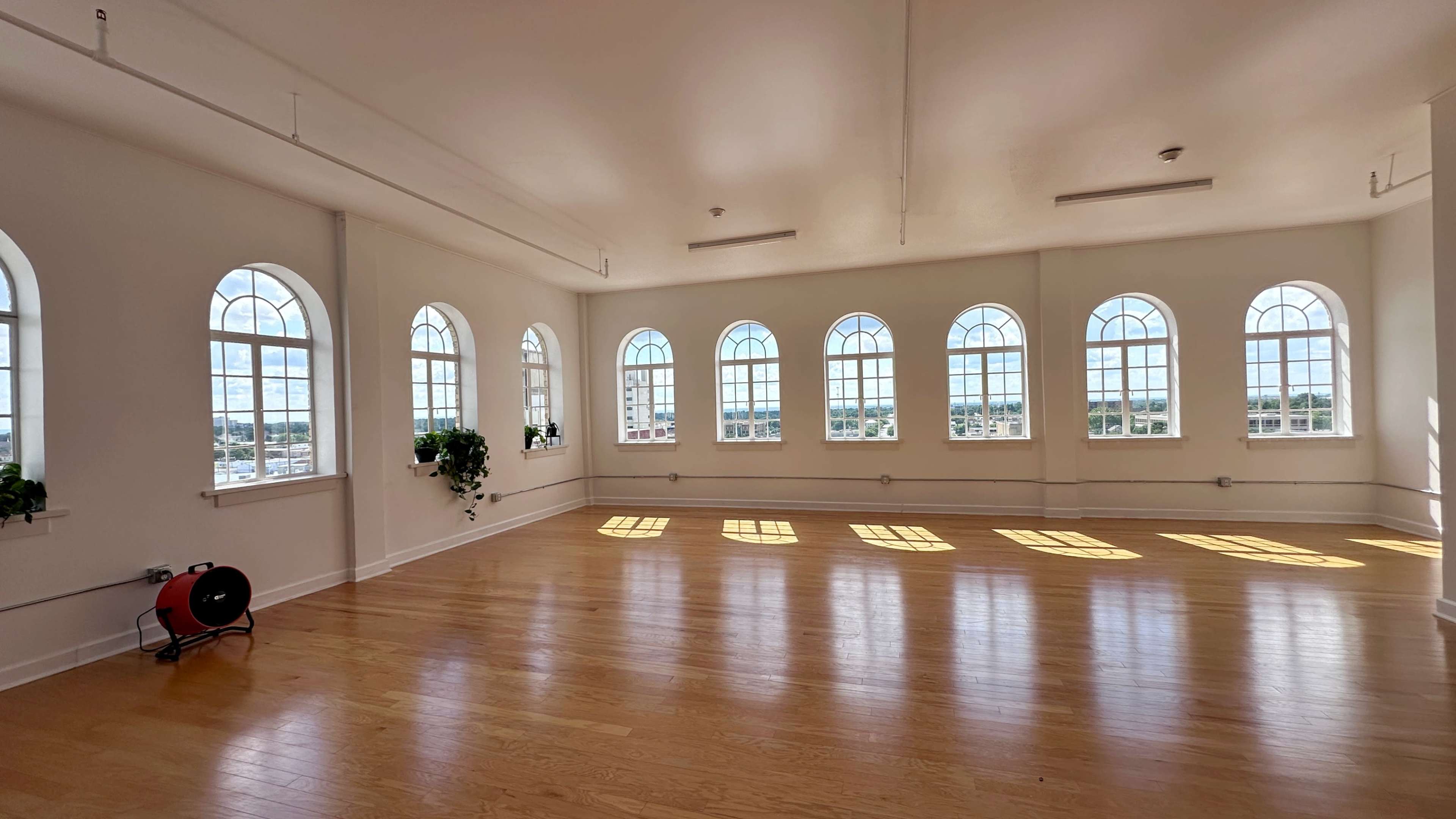 The image shows a spacious room with large arched windows that allow natural light to fill the space, highlighting the wooden floor and white walls.
