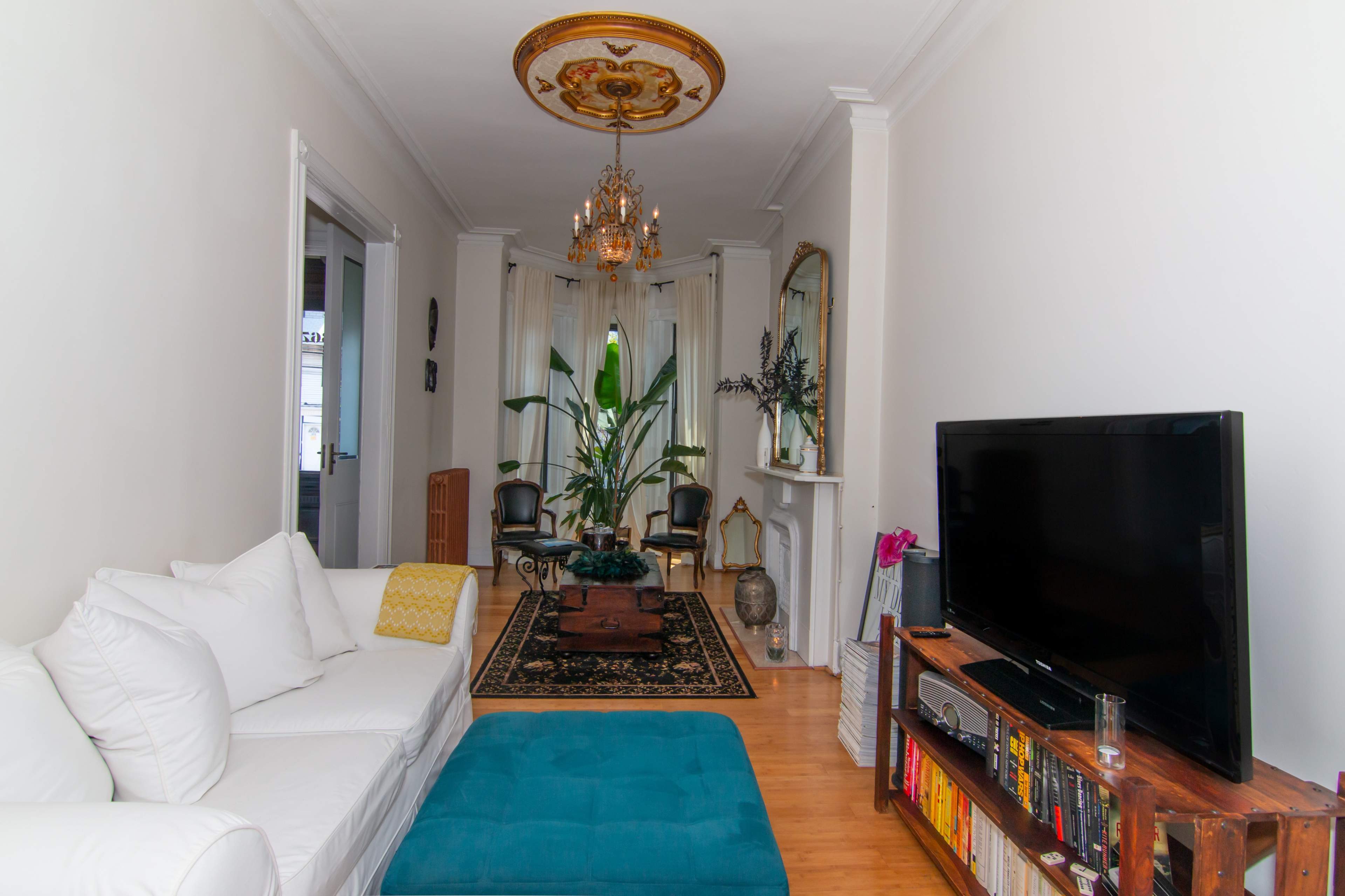 A living room featuring a white sofa, a large television, a wooden shelf with books, and ornamental plants, all set under a decorative ceiling and chandelier.