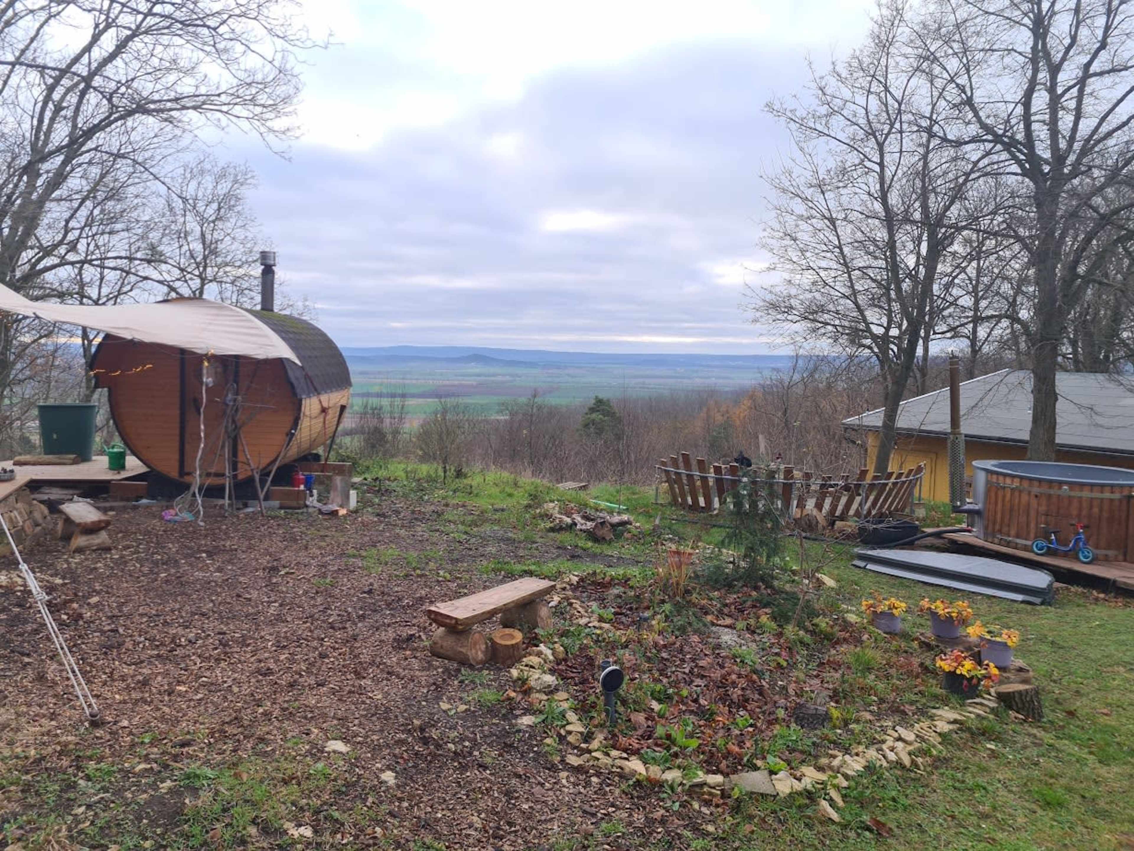 The image shows a rustic outdoor area featuring a round wooden sauna, a hot tub, a seating arrangement made of logs, and a view of rolling hills under an overcast sky.
