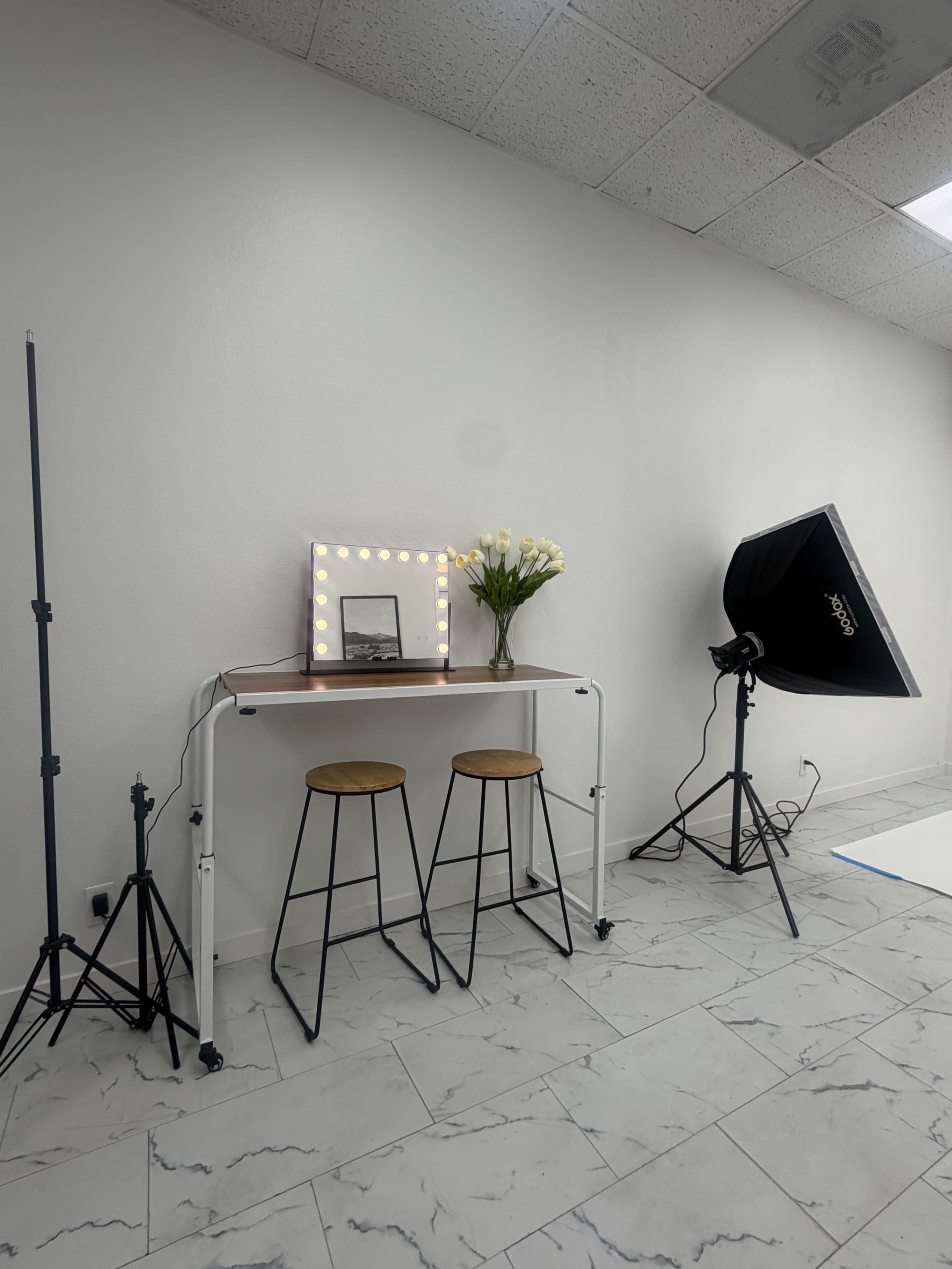 The image shows a well-lit photography studio setup featuring two high stools, a table with a mirror surrounded by lights, and a bouquet of flowers.