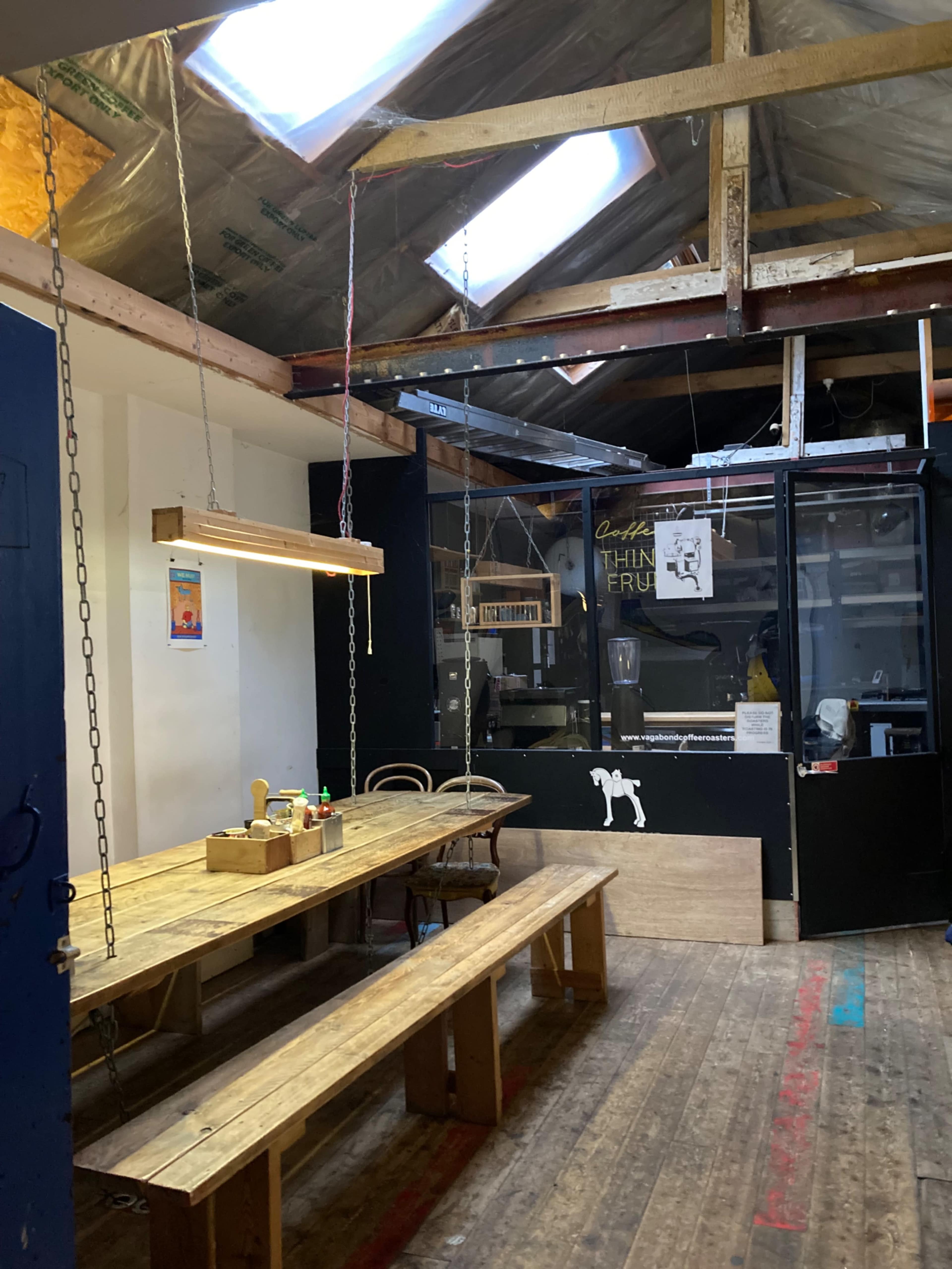 The image shows a rustic interior with a wooden table and benches, overhead lighting, and a kitchen area visible in the background.