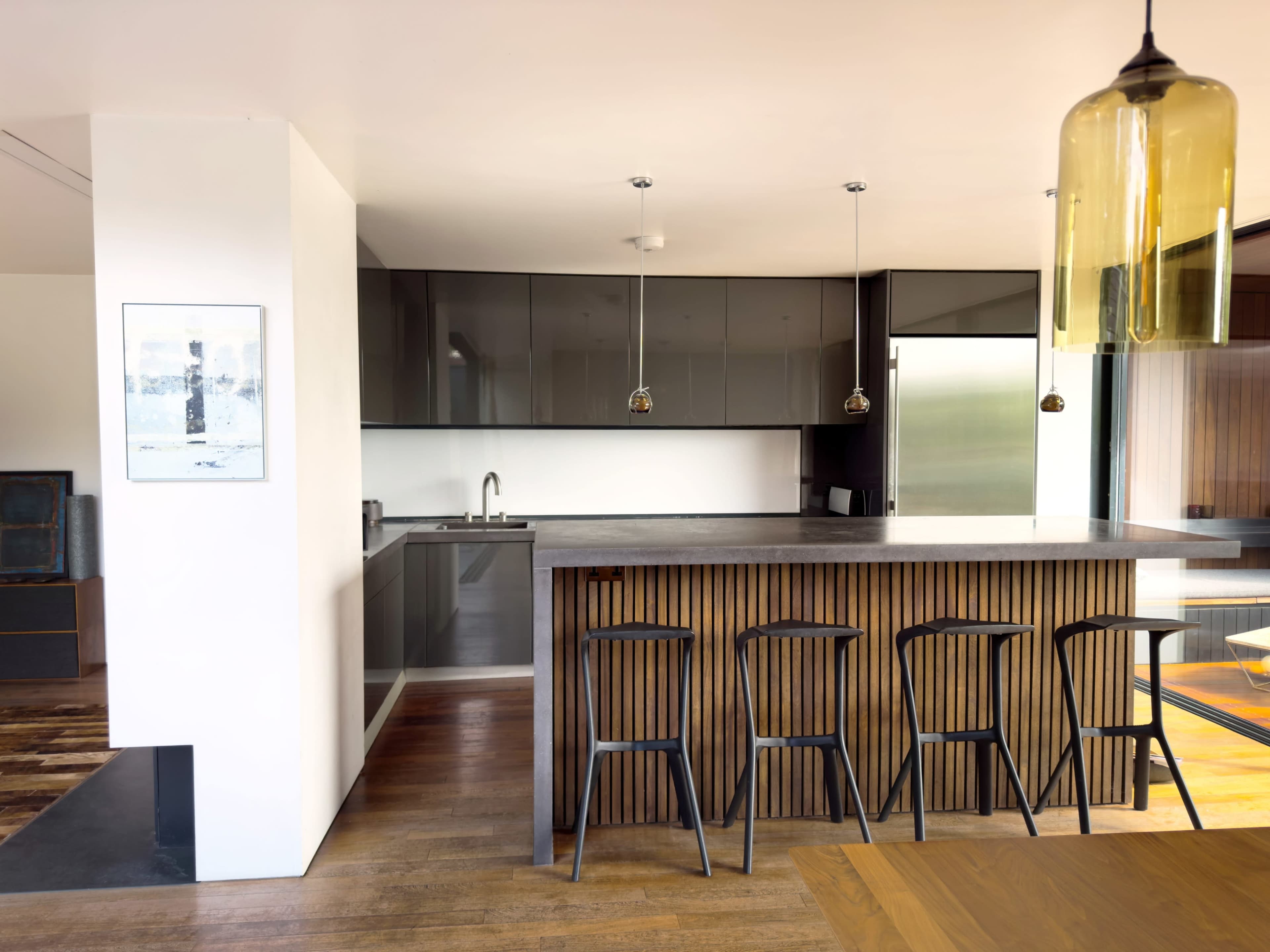 A modern kitchen features sleek dark cabinetry, a spacious island with wooden accents, and minimalist black bar stools.