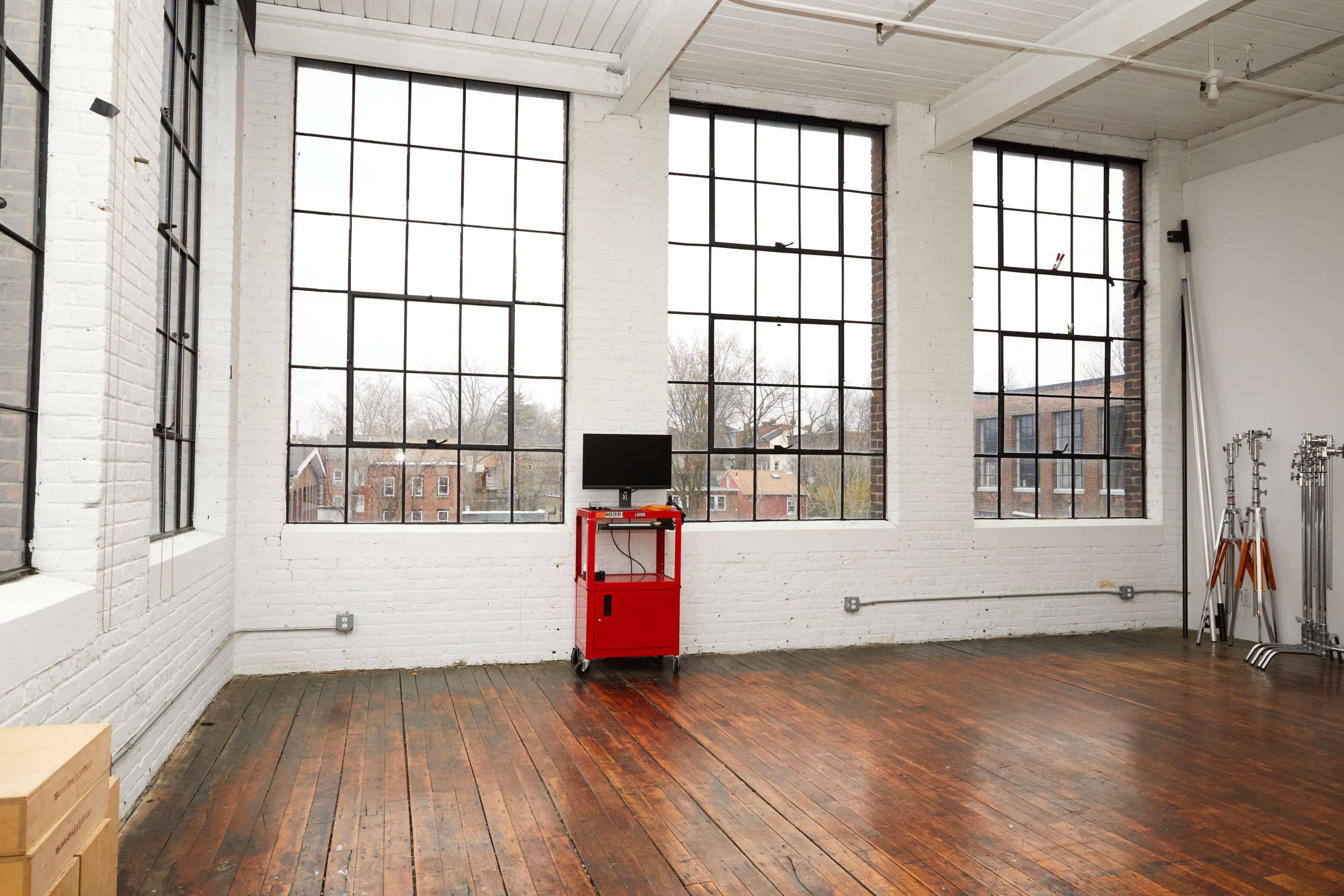 An empty room with large windows, wooden floors, and a red cart with a monitor positioned in front of the windows.