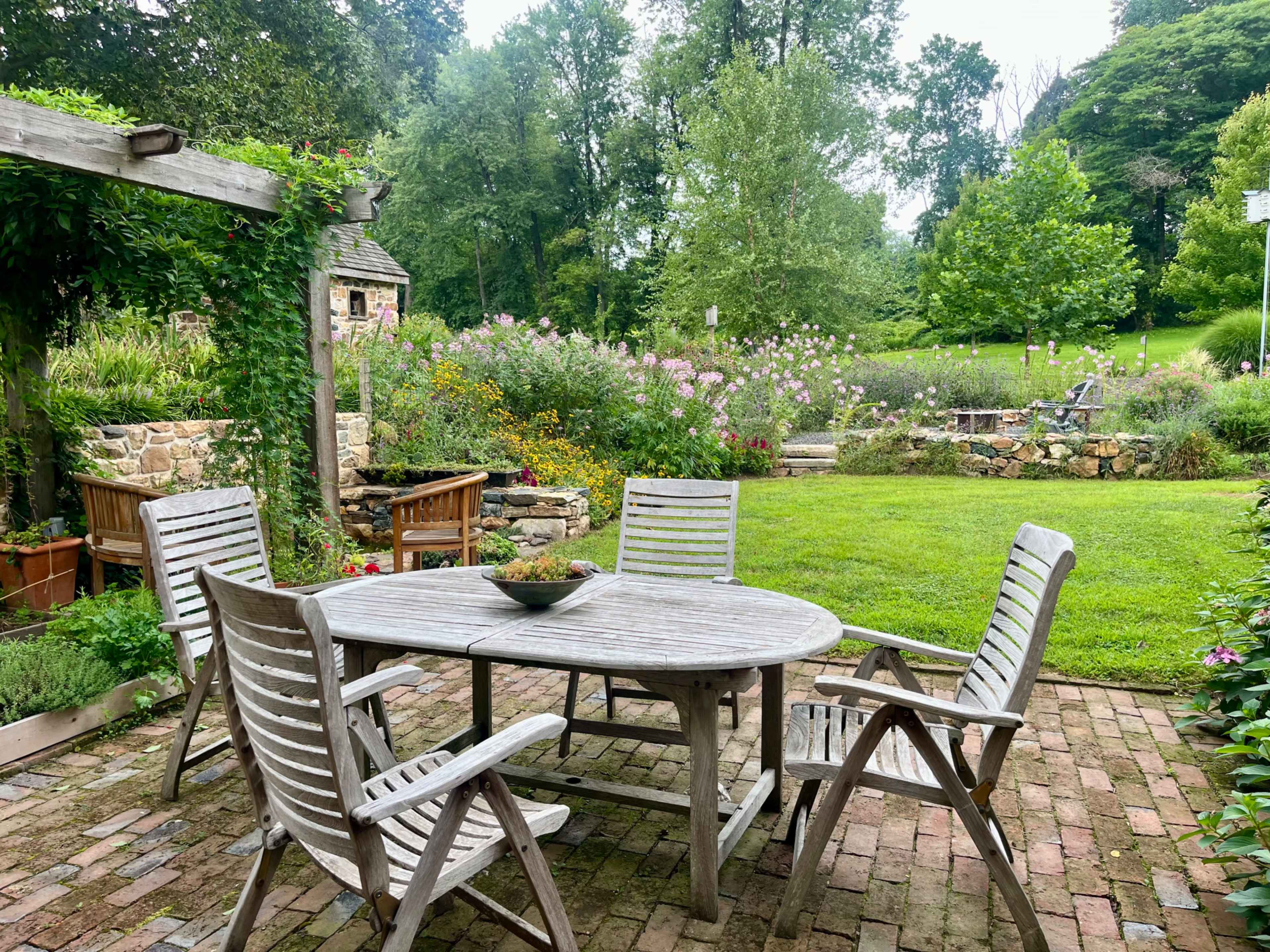 A rustic outdoor dining area features a wooden table surrounded by five chairs, set on a brick patio amidst lush greenery and colorful flowers.