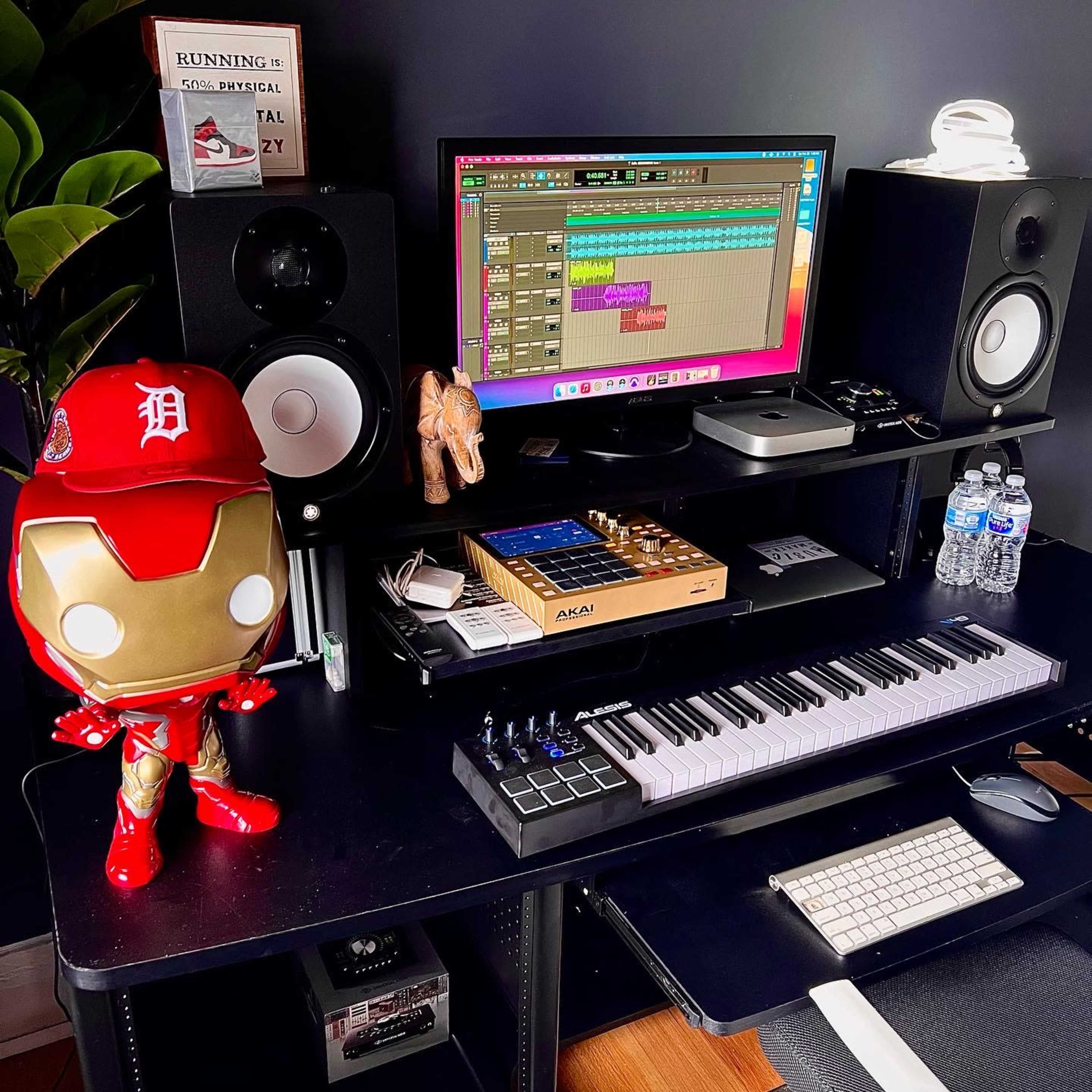 A modern music production setup featuring a computer with audio software, studio monitors, an Akai MIDI keyboard, a toy Iron Man figure, and a few bottles of water on a black desk.