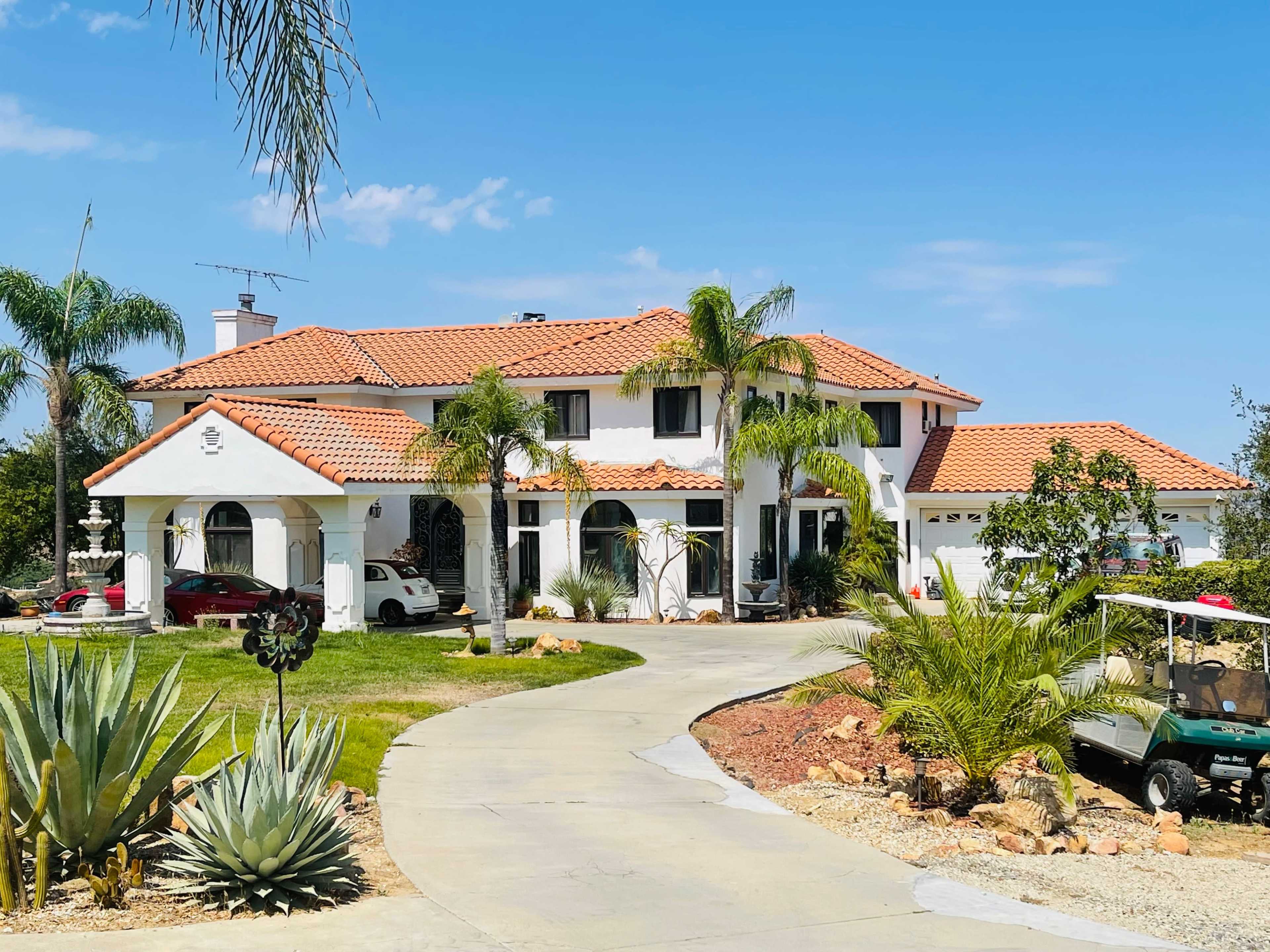 A large white house with a red tile roof, surrounded by palm trees and a circular driveway.