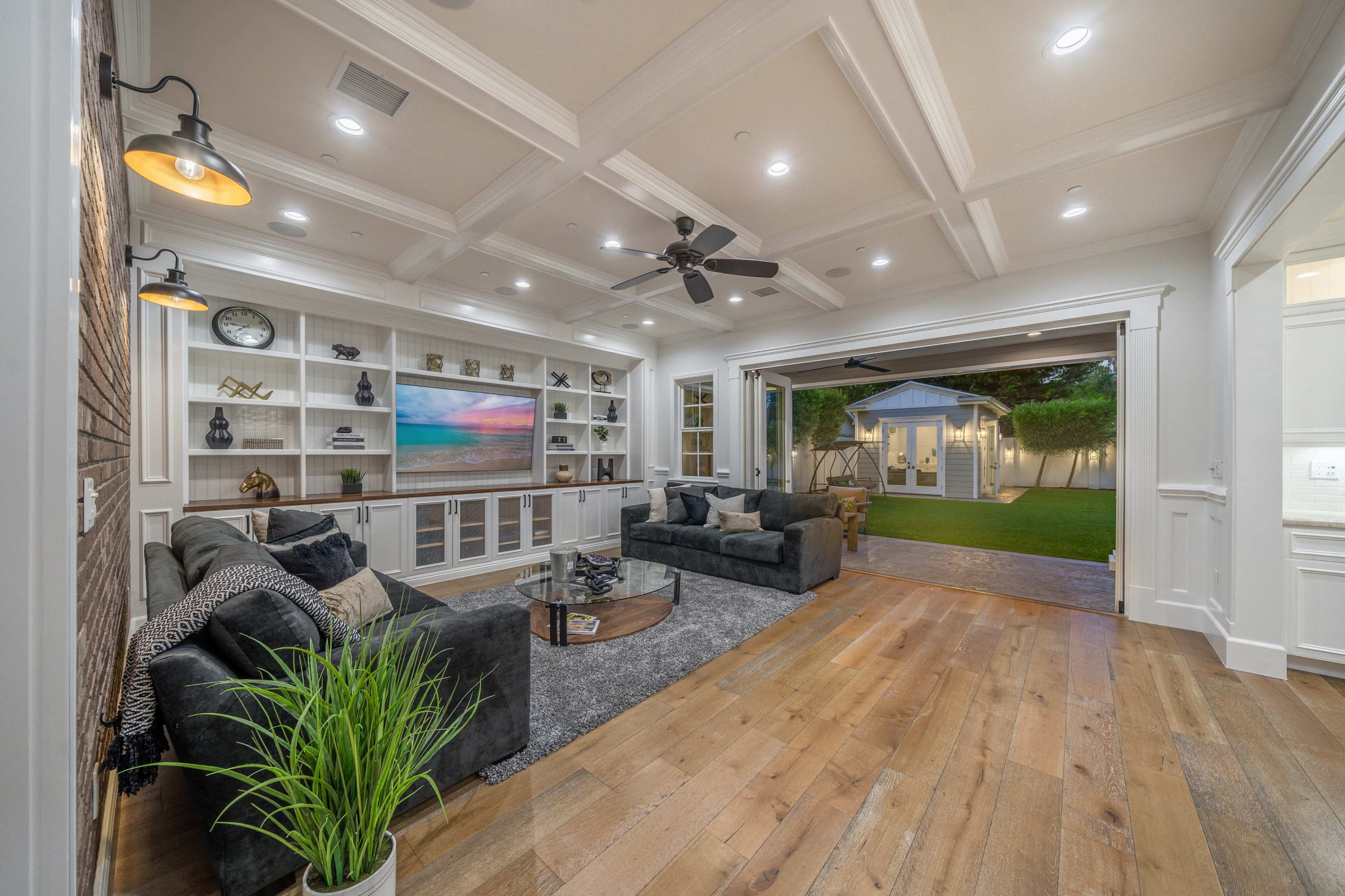 A modern living room with a coffered ceiling, a display cabinet, two sofas, and an open wall that leads to a backyard with a small shed.