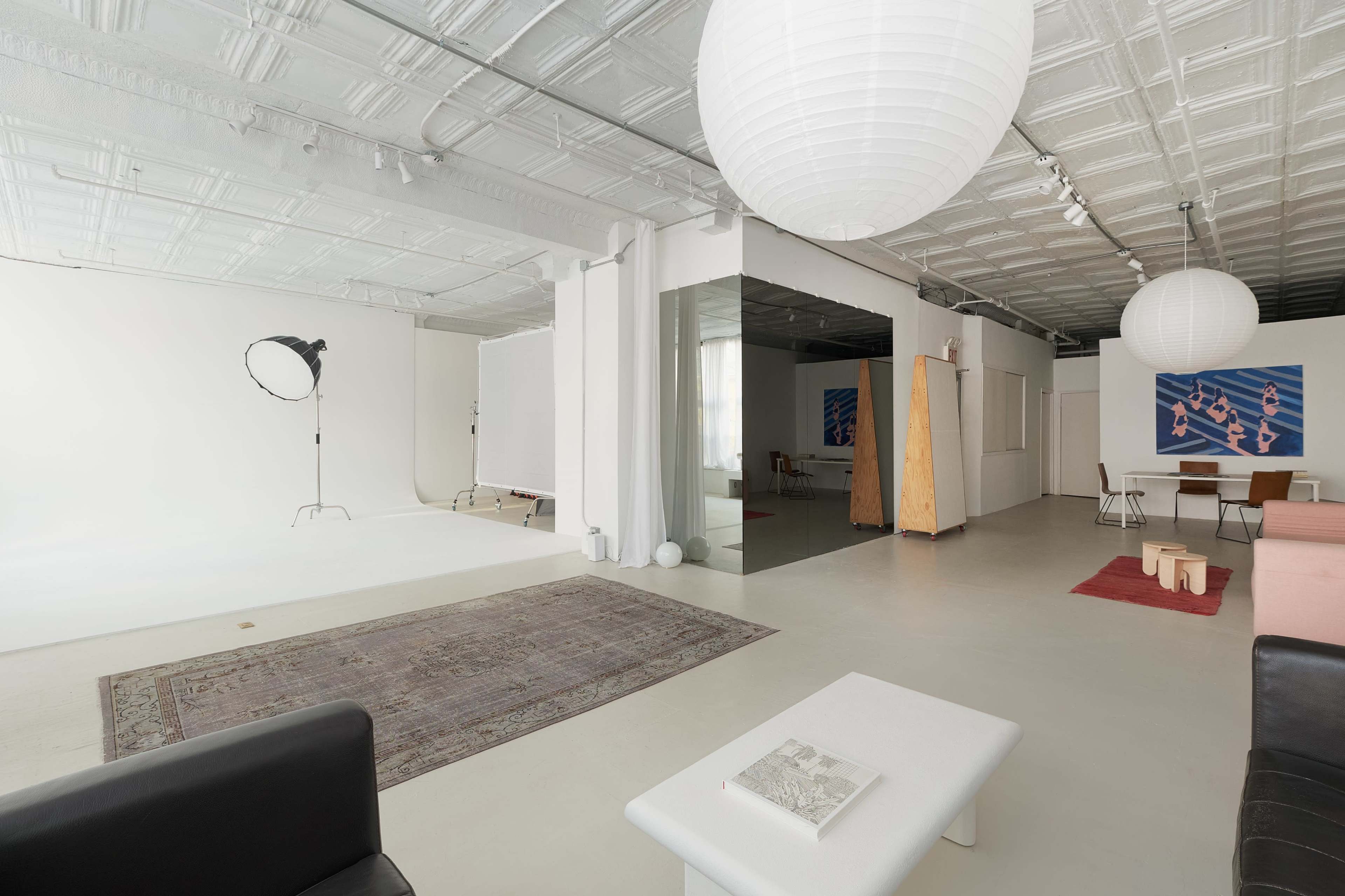 The image shows a spacious studio with white walls, a patterned rug, and soft lighting fixtures hanging from the ceiling, along with various furniture pieces and artistic displays.