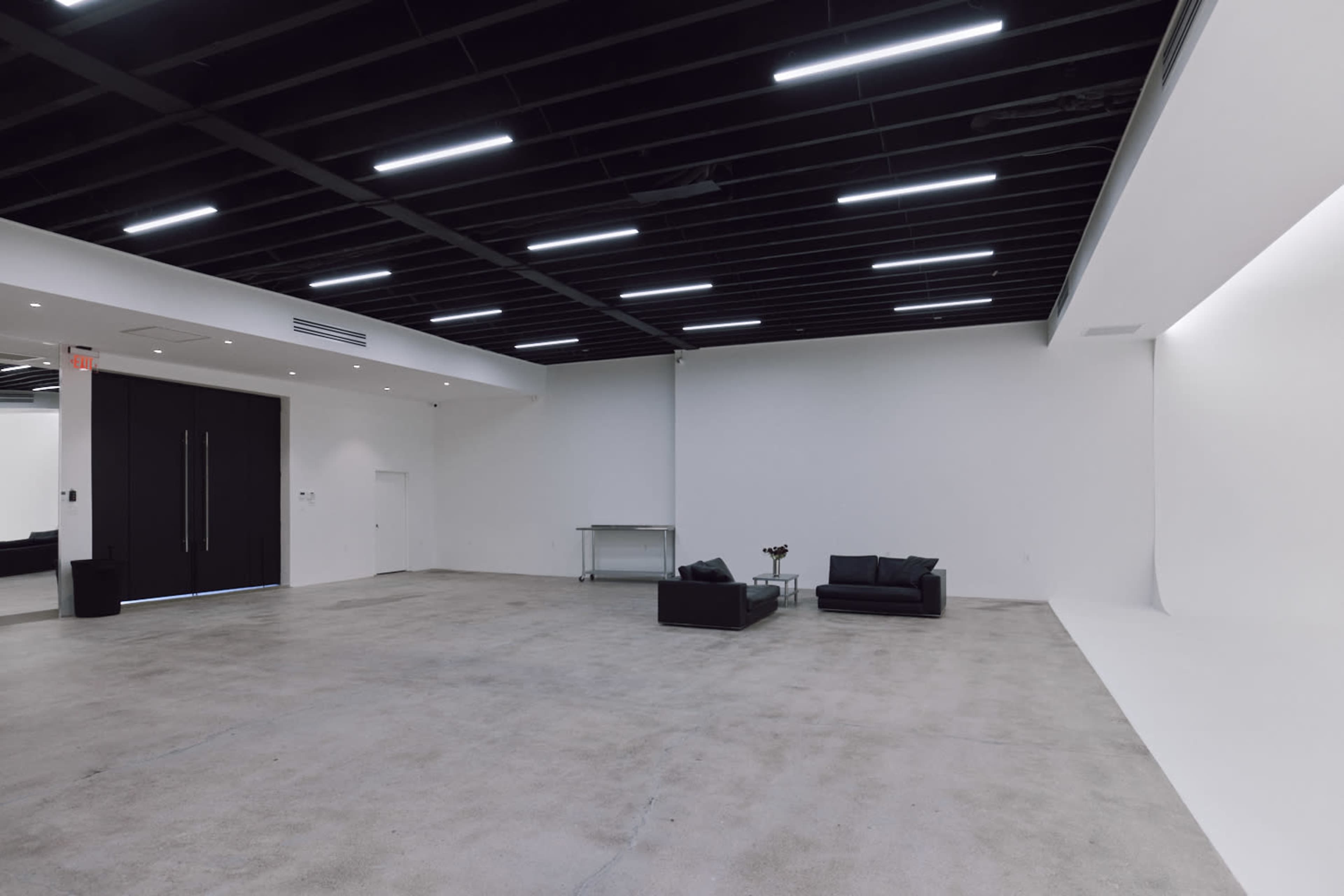 The image shows a spacious, minimalistic room with a concrete floor, black ceiling beams, and a black couch positioned on one side.