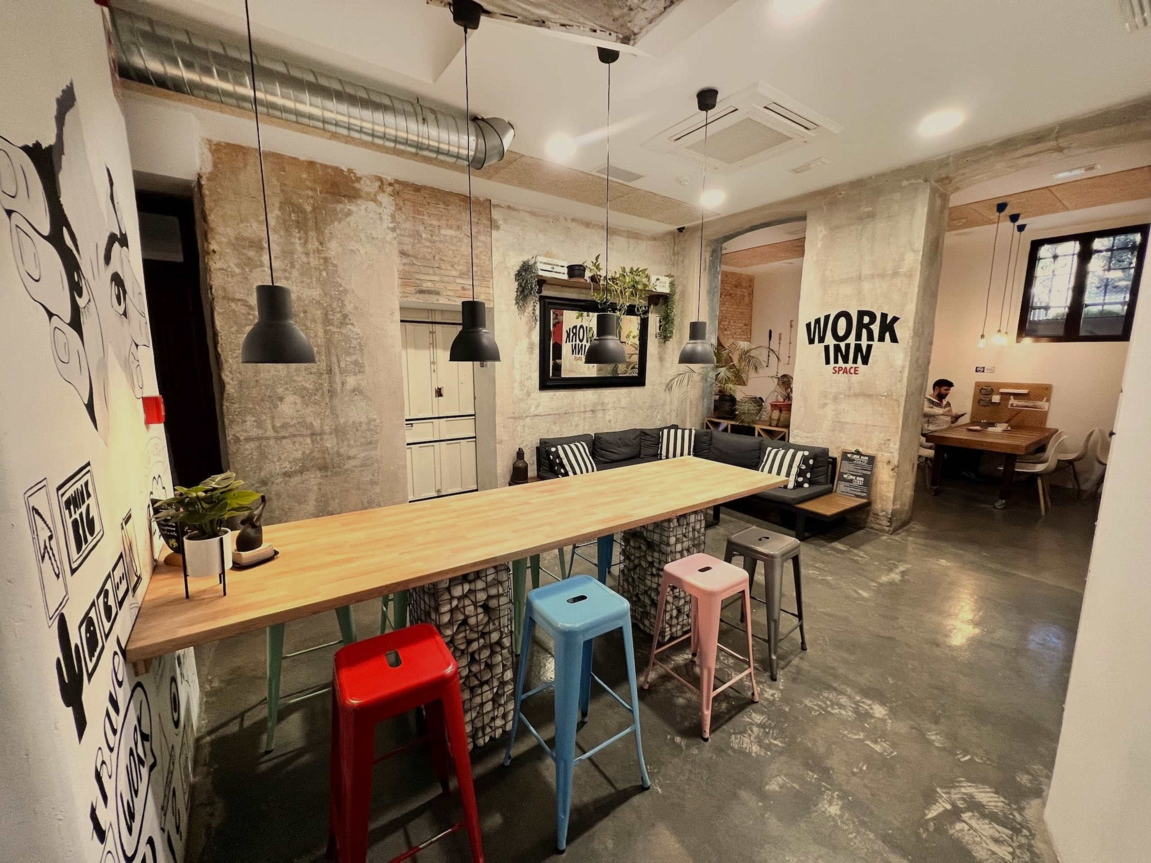 The image shows a modern coworking space featuring a long wooden table with colorful stools, concrete walls, and a relaxed seating area.