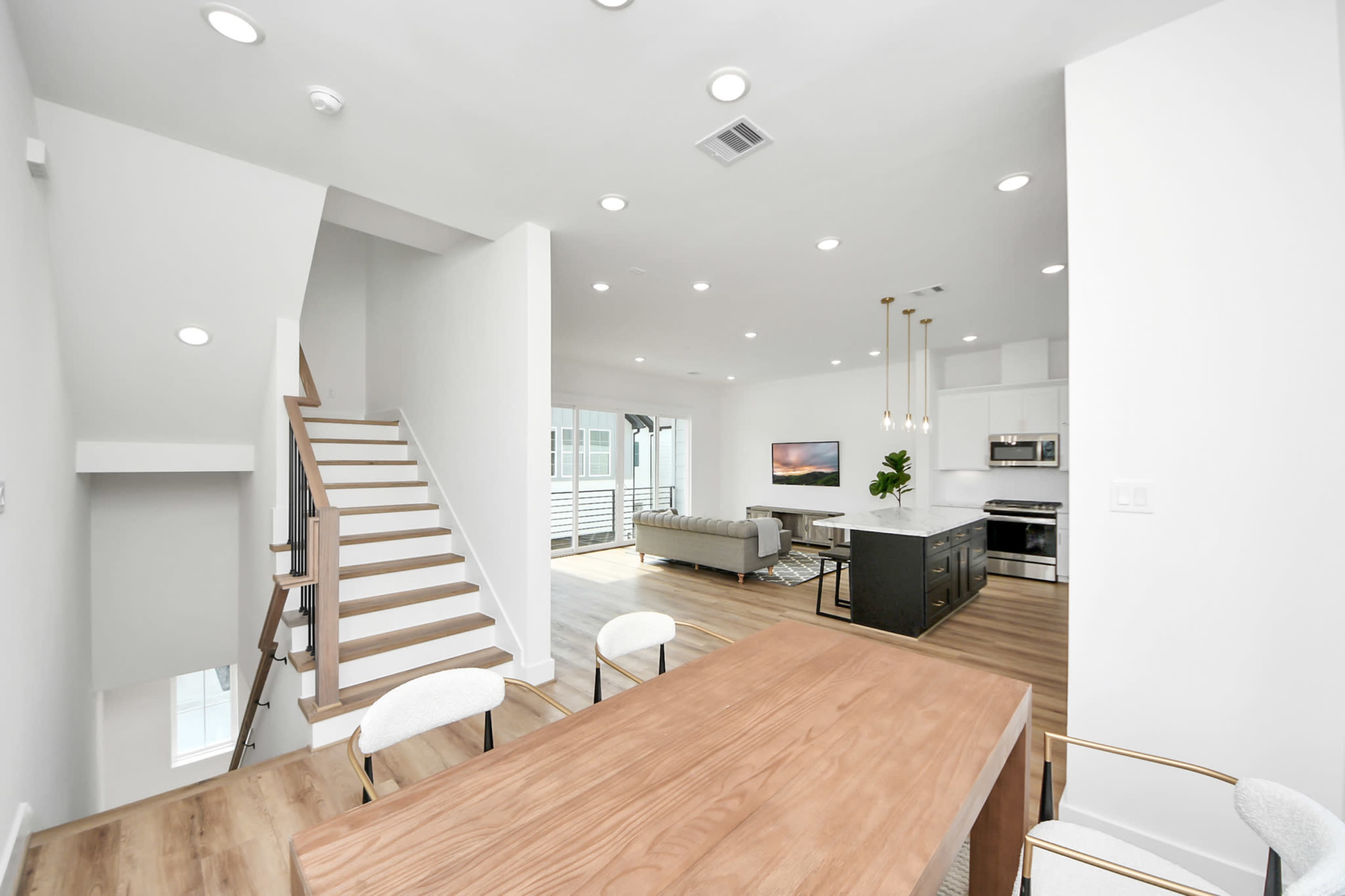 The image shows a modern, open-concept living area and dining space with a staircase, kitchen island, and large windows.