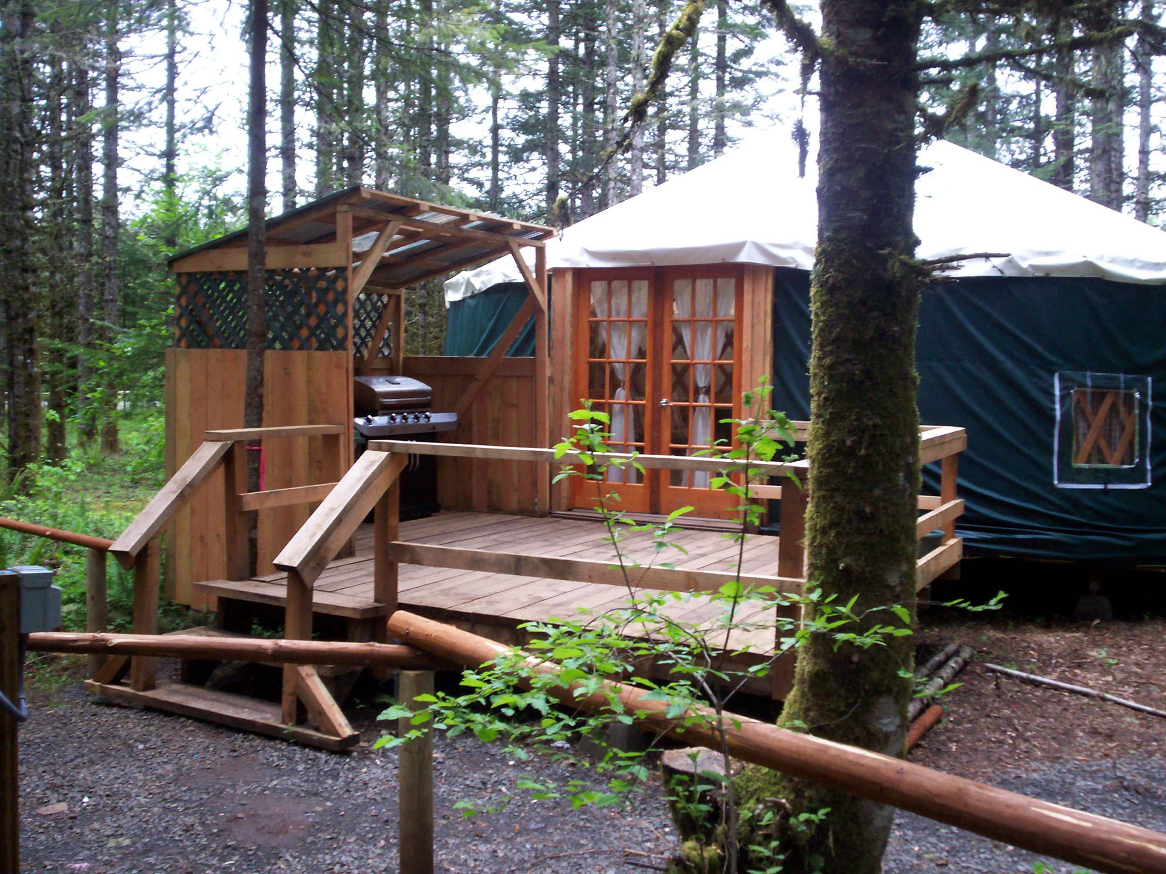 A wooden deck with a barbecue grill beside a yurt, nestled in a forested area.