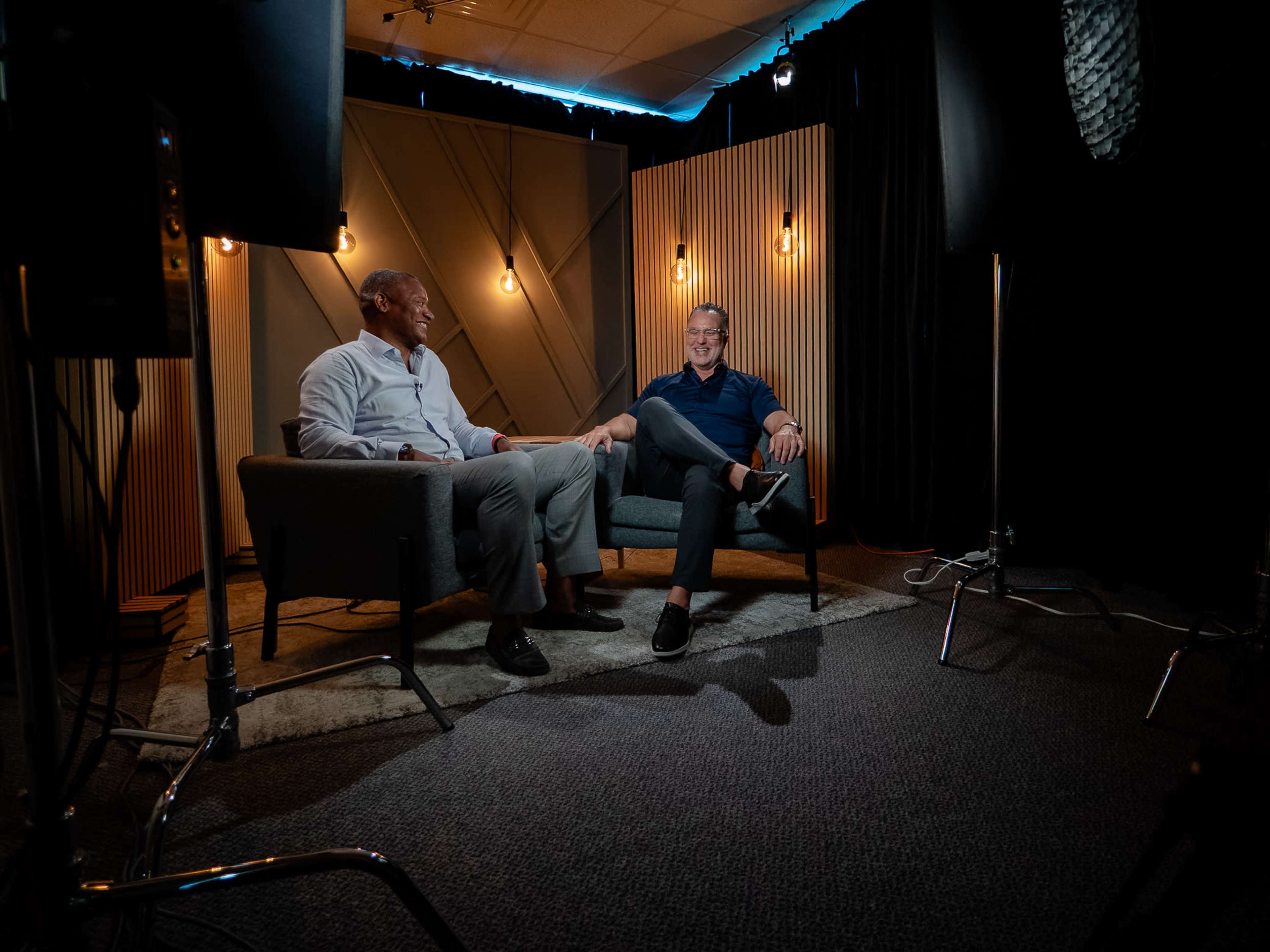 Two men are seated on a couch in a well-lit interview setting with decorative acoustic panels in the background.