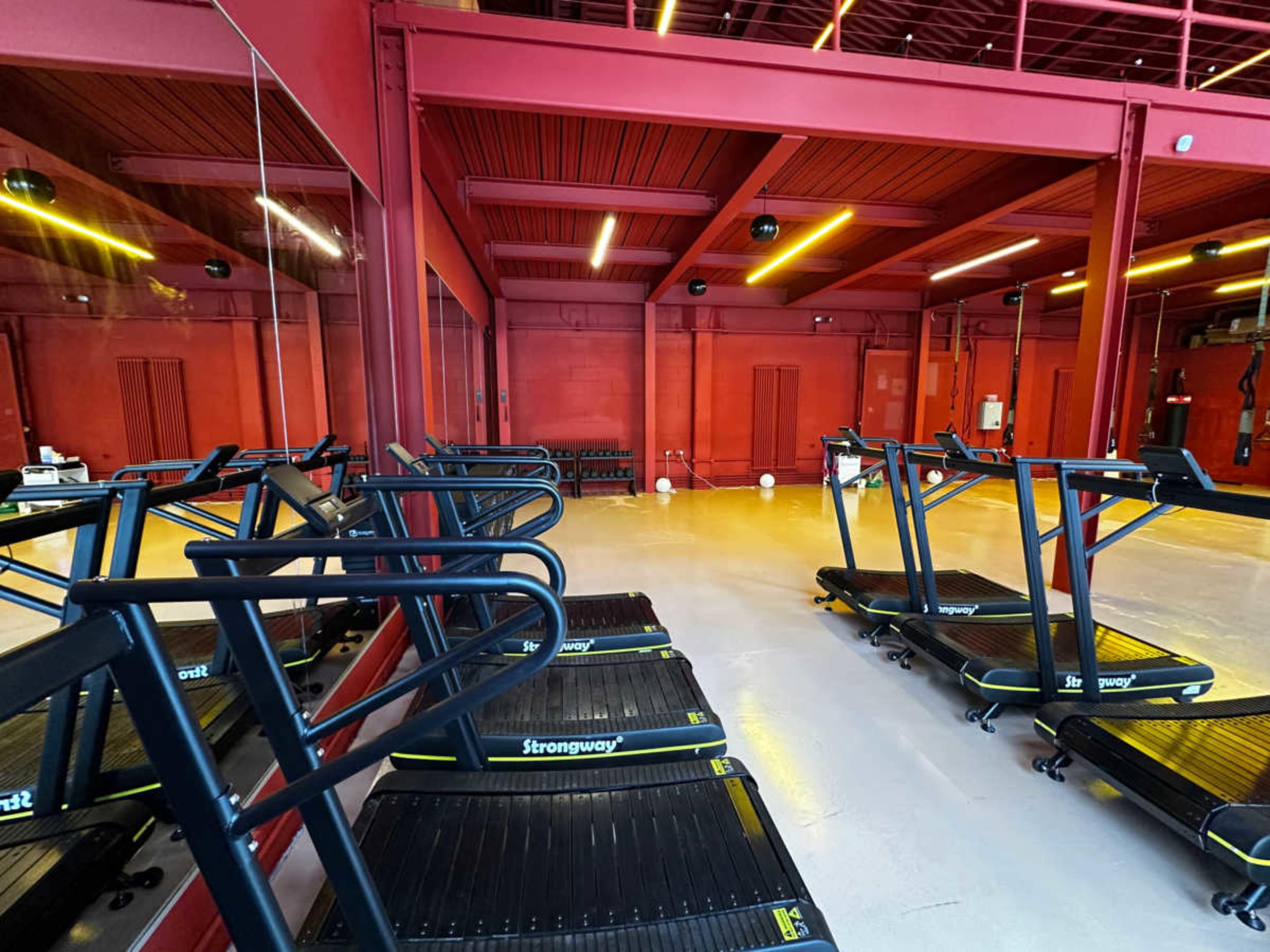 The image shows a fitness studio with several treadmill machines arranged neatly against a red wall.