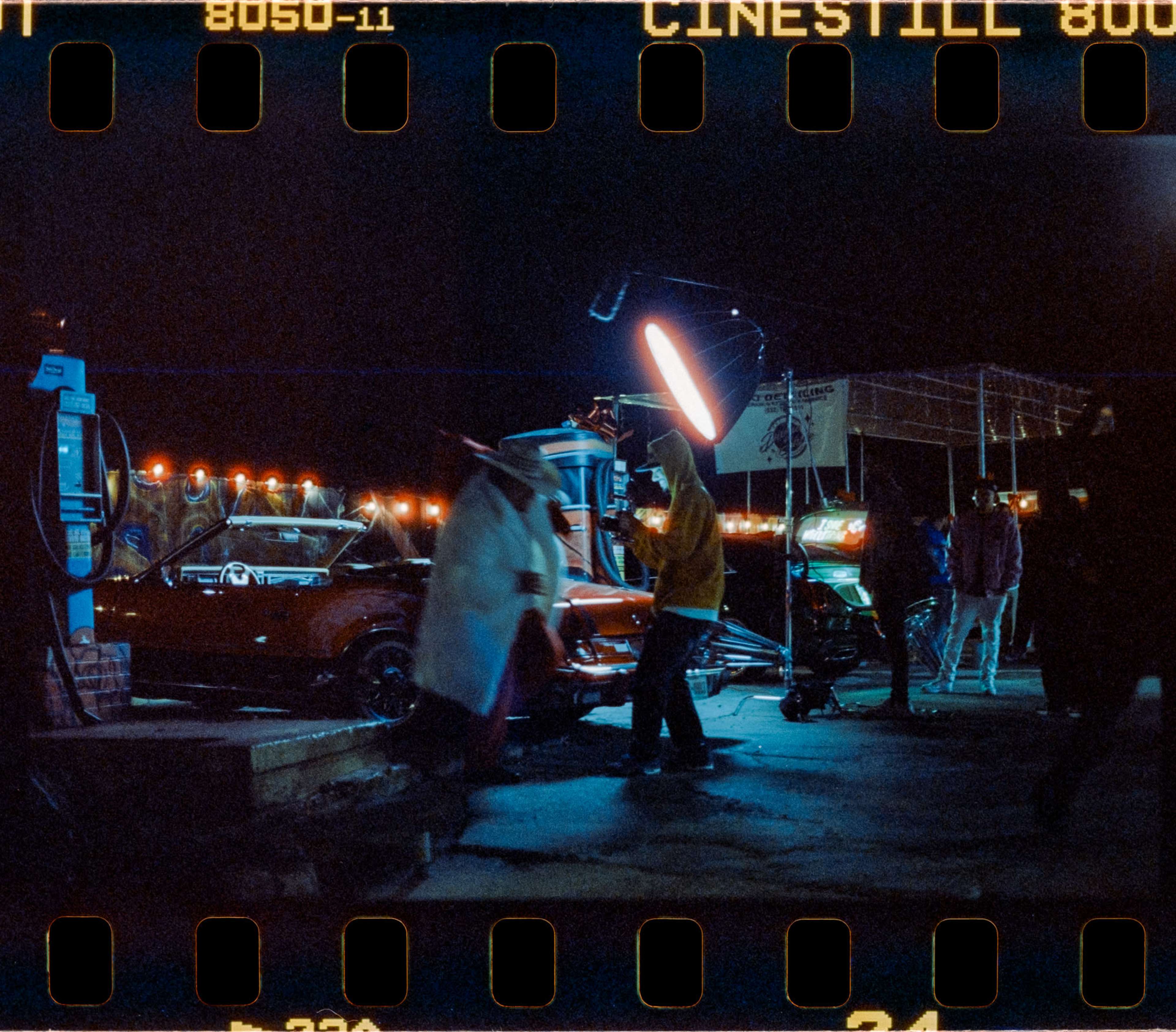 The scene depicts a nighttime shooting location with cars, crew members, and lighting equipment set up for a film or photo shoot.