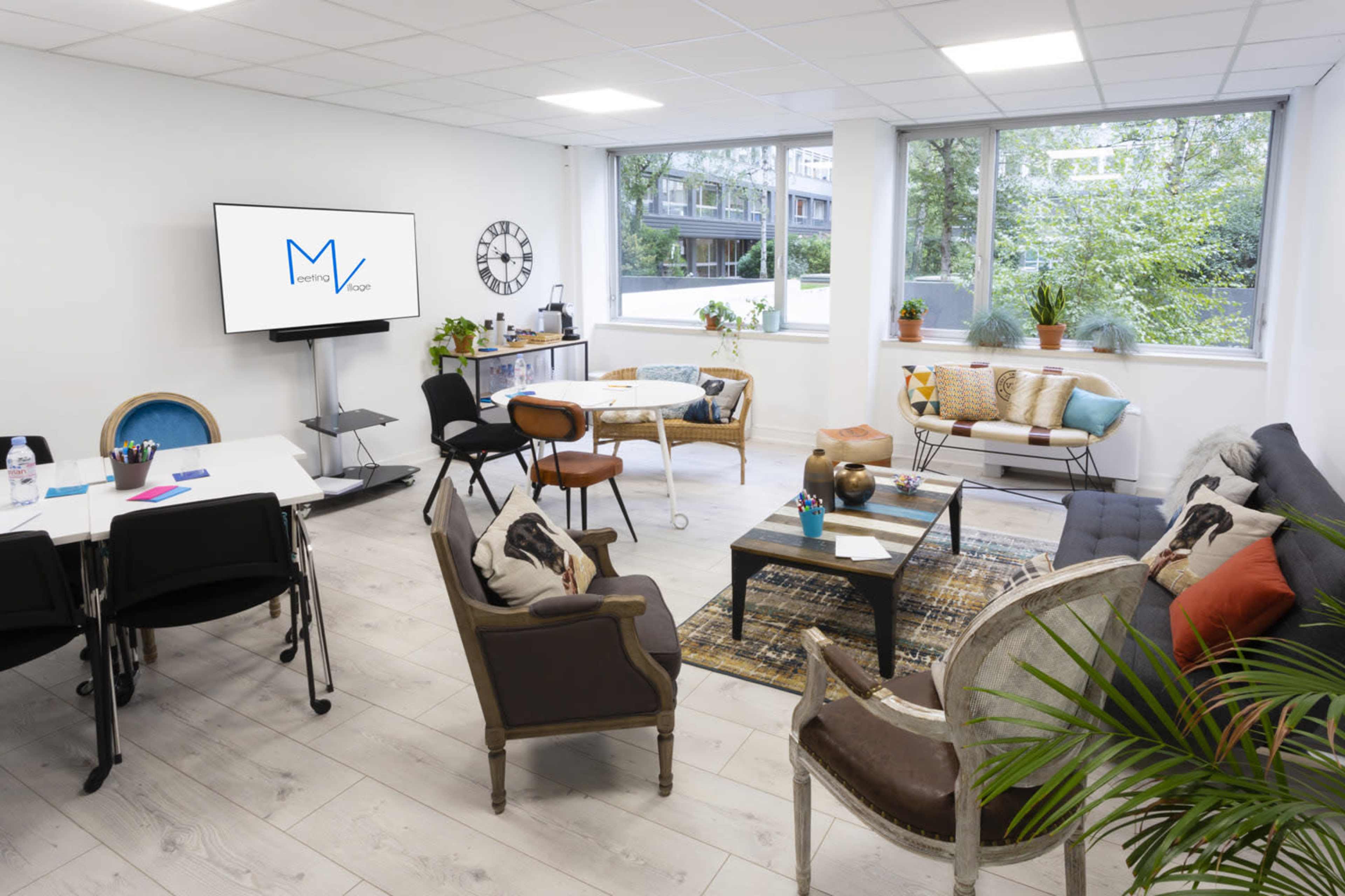 The image shows a bright office space featuring a meeting area with a large screen, several seating arrangements, and windows that provide views of greenery outside.