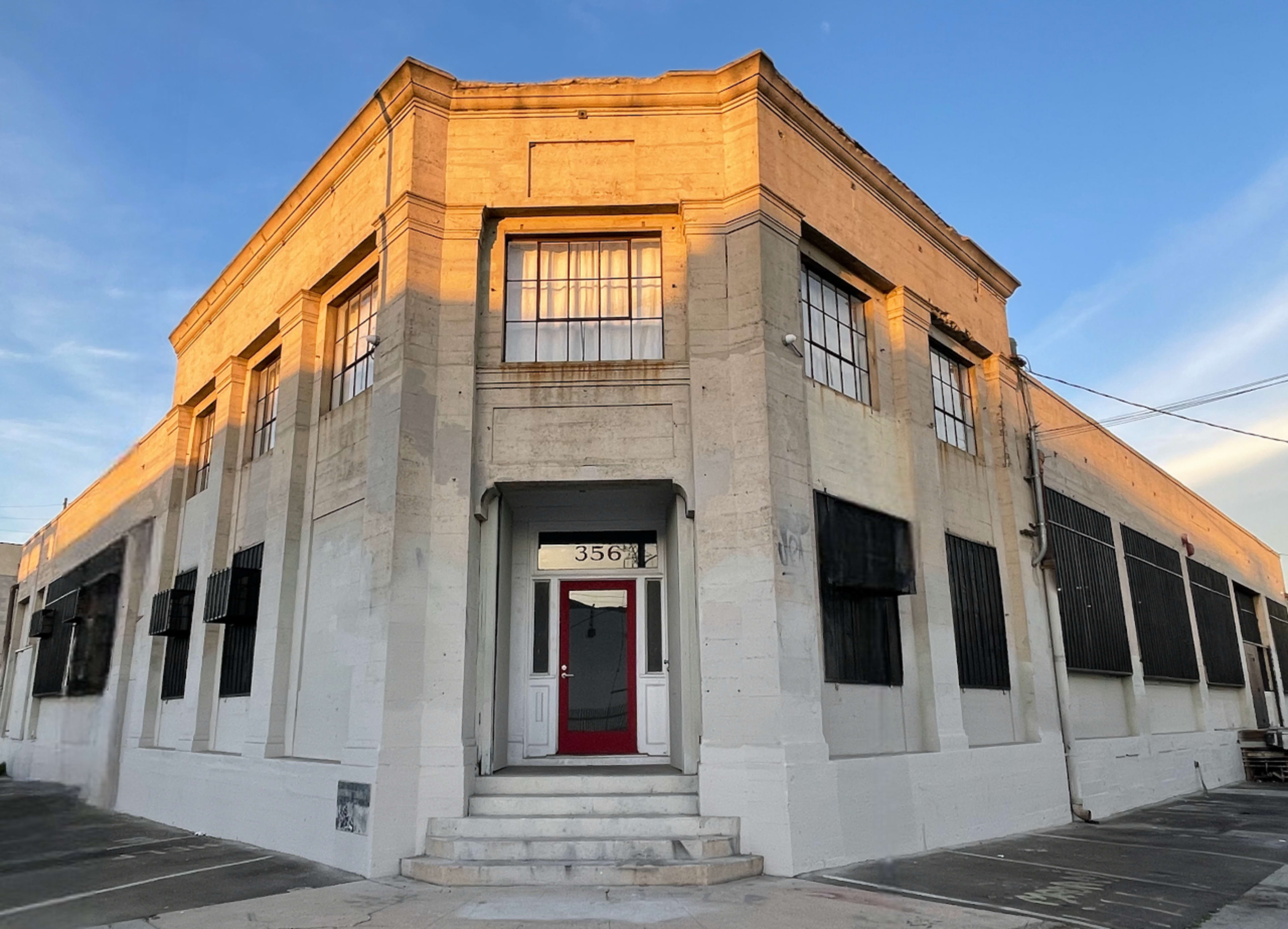 The image shows a large, corner building with a white exterior, multiple windows, and a red door prominently featured at the entrance.