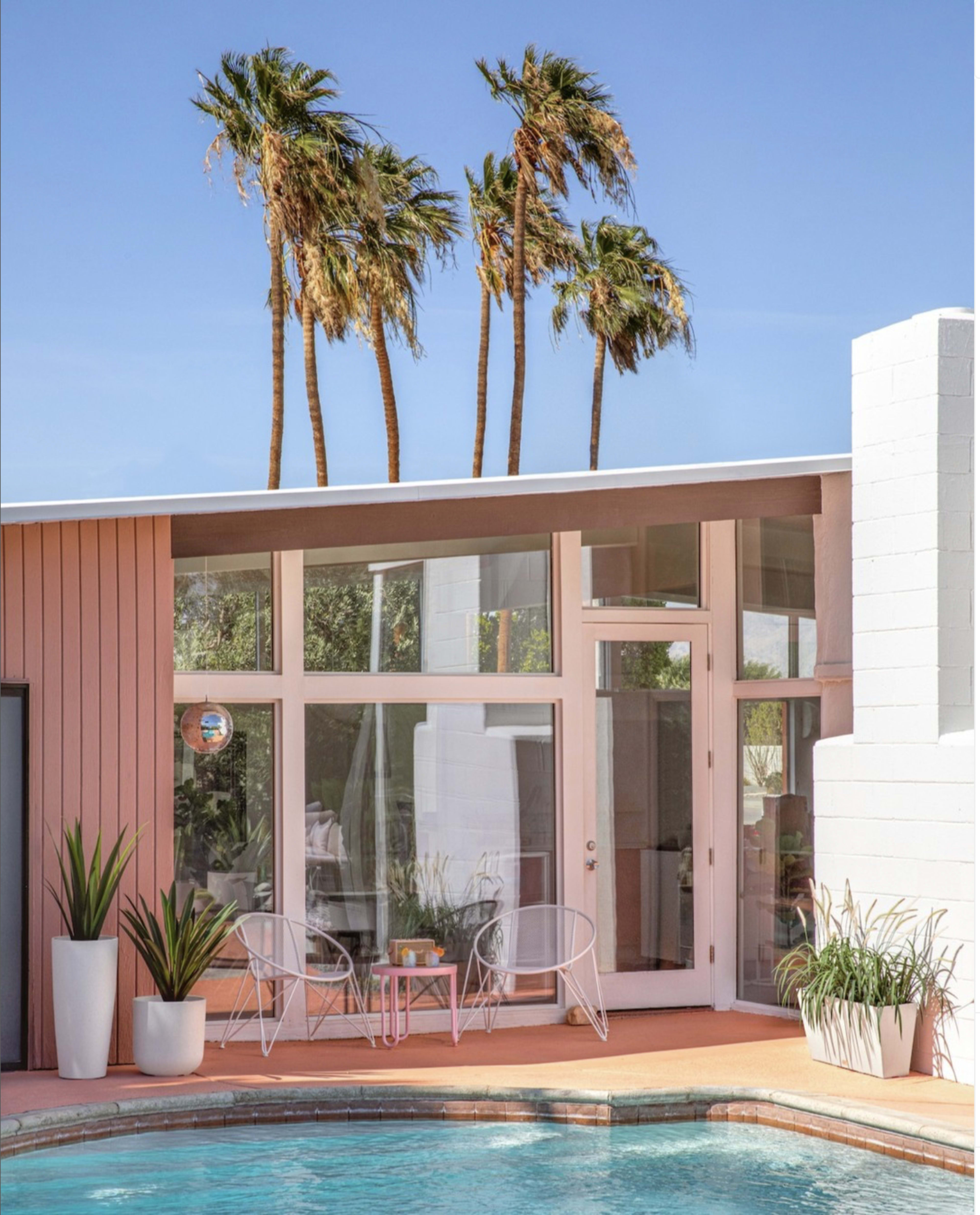 A modern house with large windows overlooks a swimming pool and is surrounded by tall palm trees.