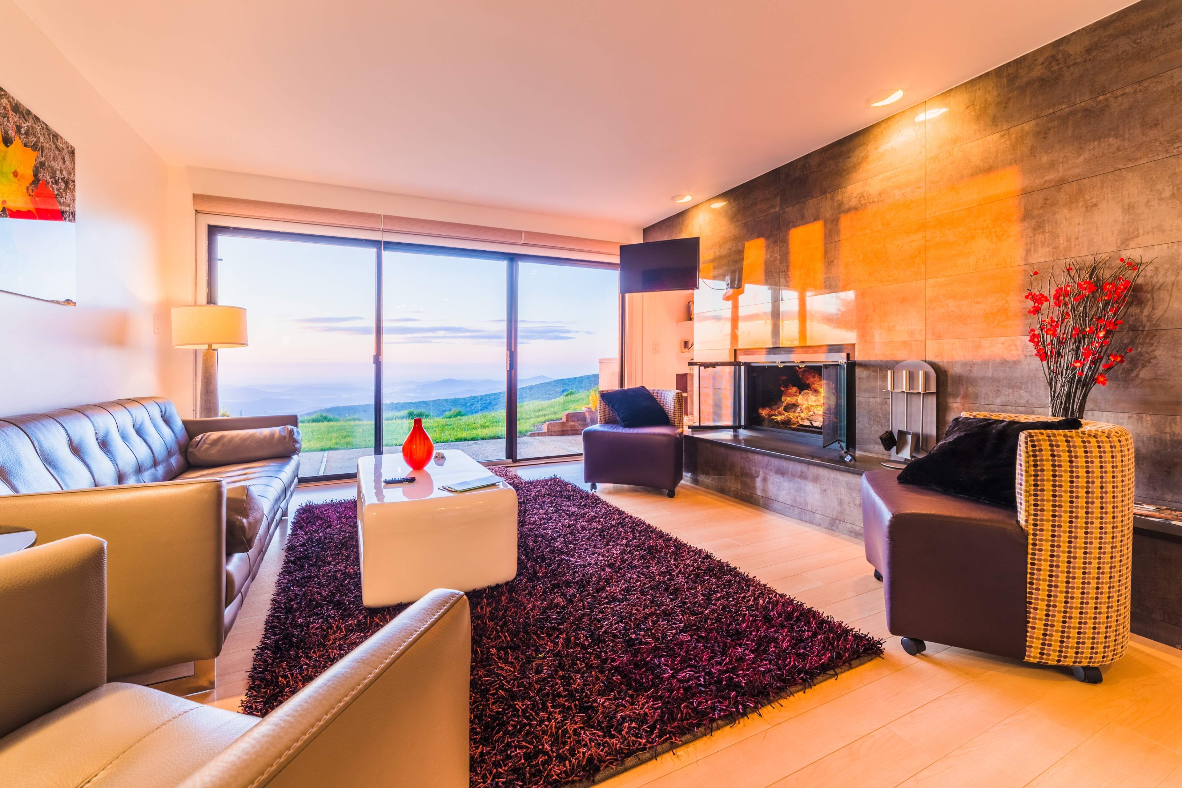A modern living room with a fireplace, large windows, and plush seating arranged on a textured rug.