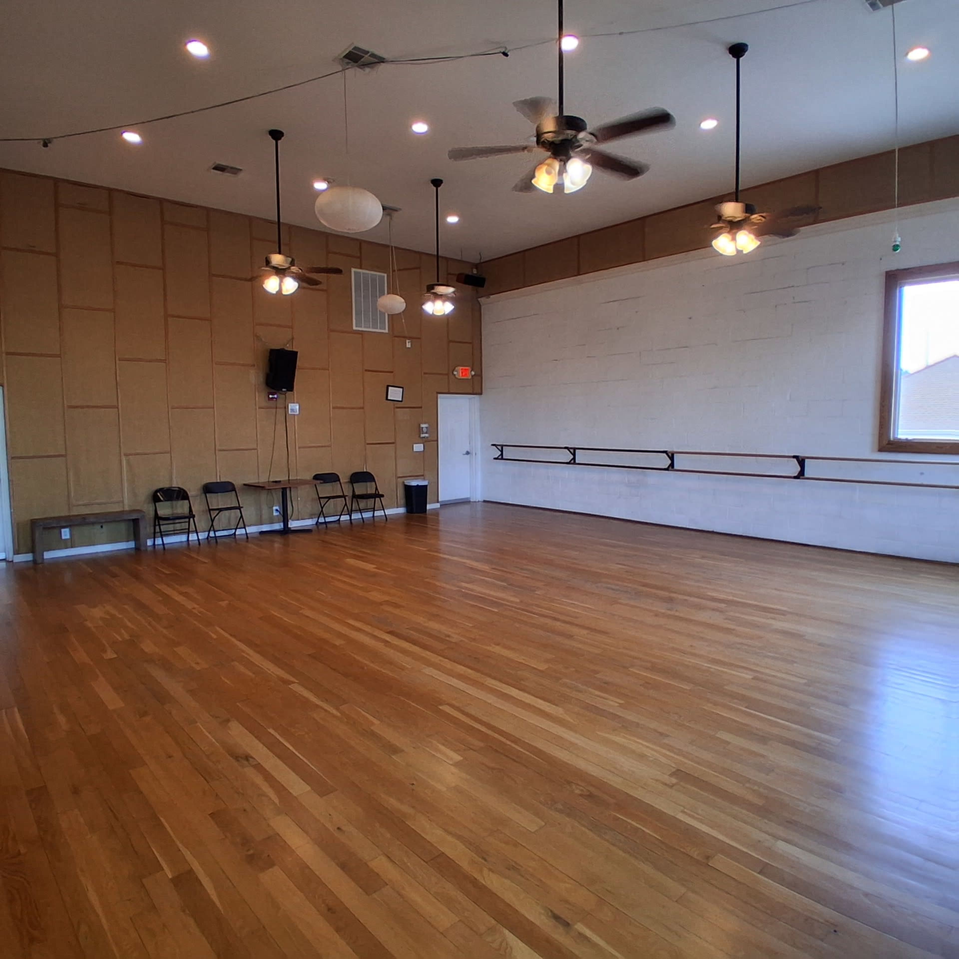 The image shows a spacious dance studio with wooden floors, several ceiling fans, and a row of black chairs along one wall.