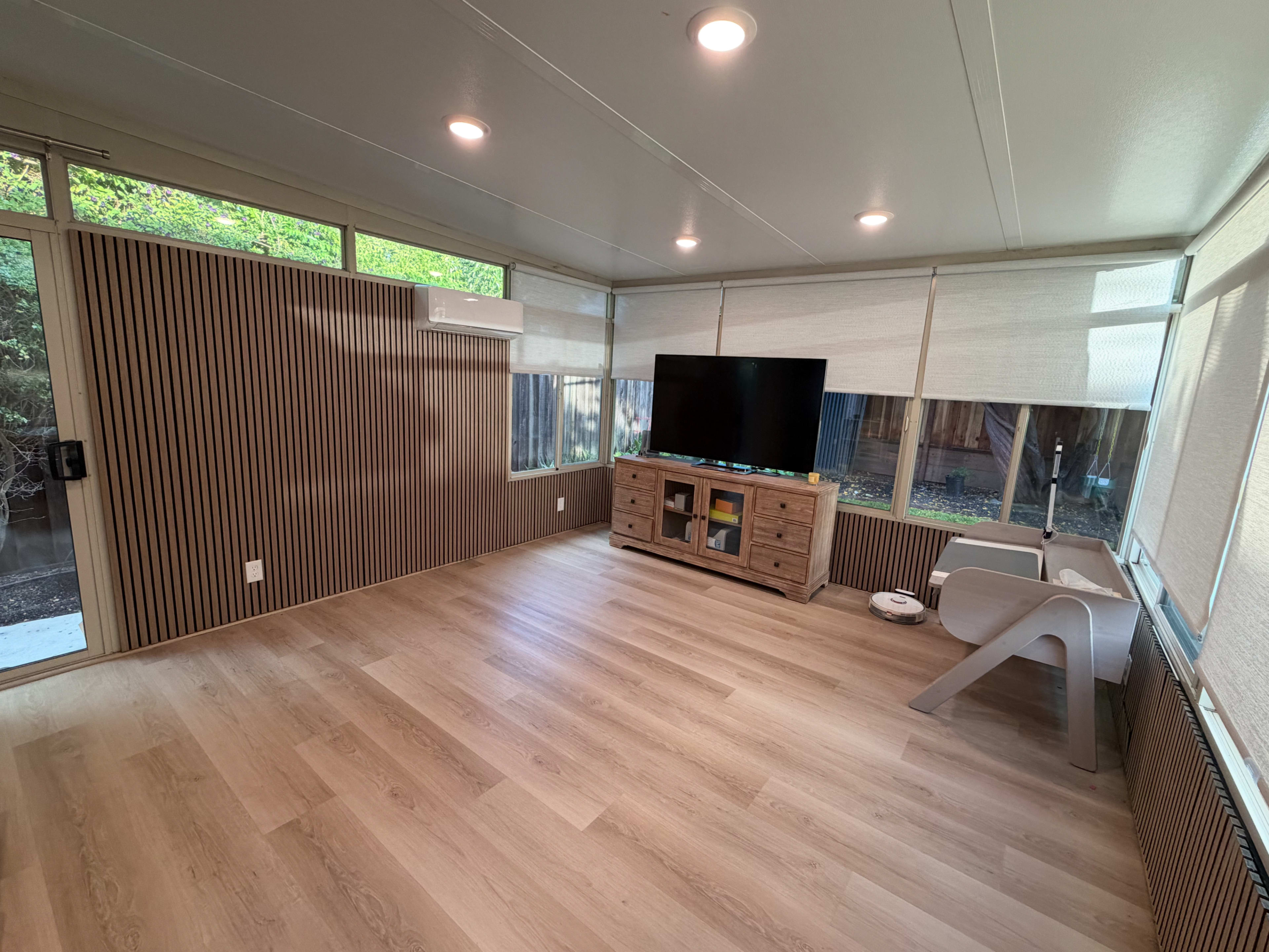 The image shows a spacious, modern interior with light wood flooring, a TV mounted on a wall, and minimal furniture including a side table and a console.