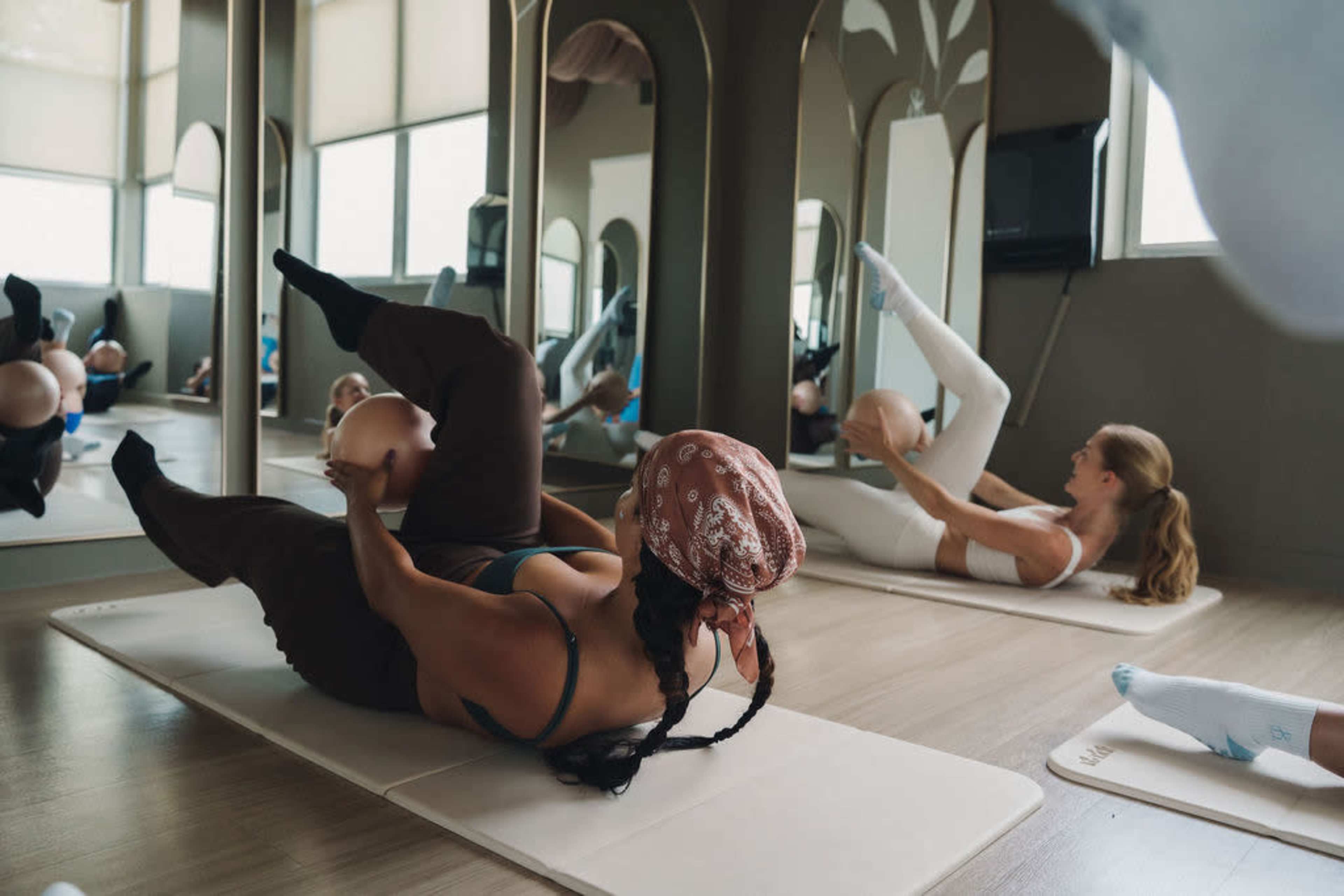 Two women are practicing pilates in a studio with mirrors, each holding a small exercise ball while lying on exercise mats.